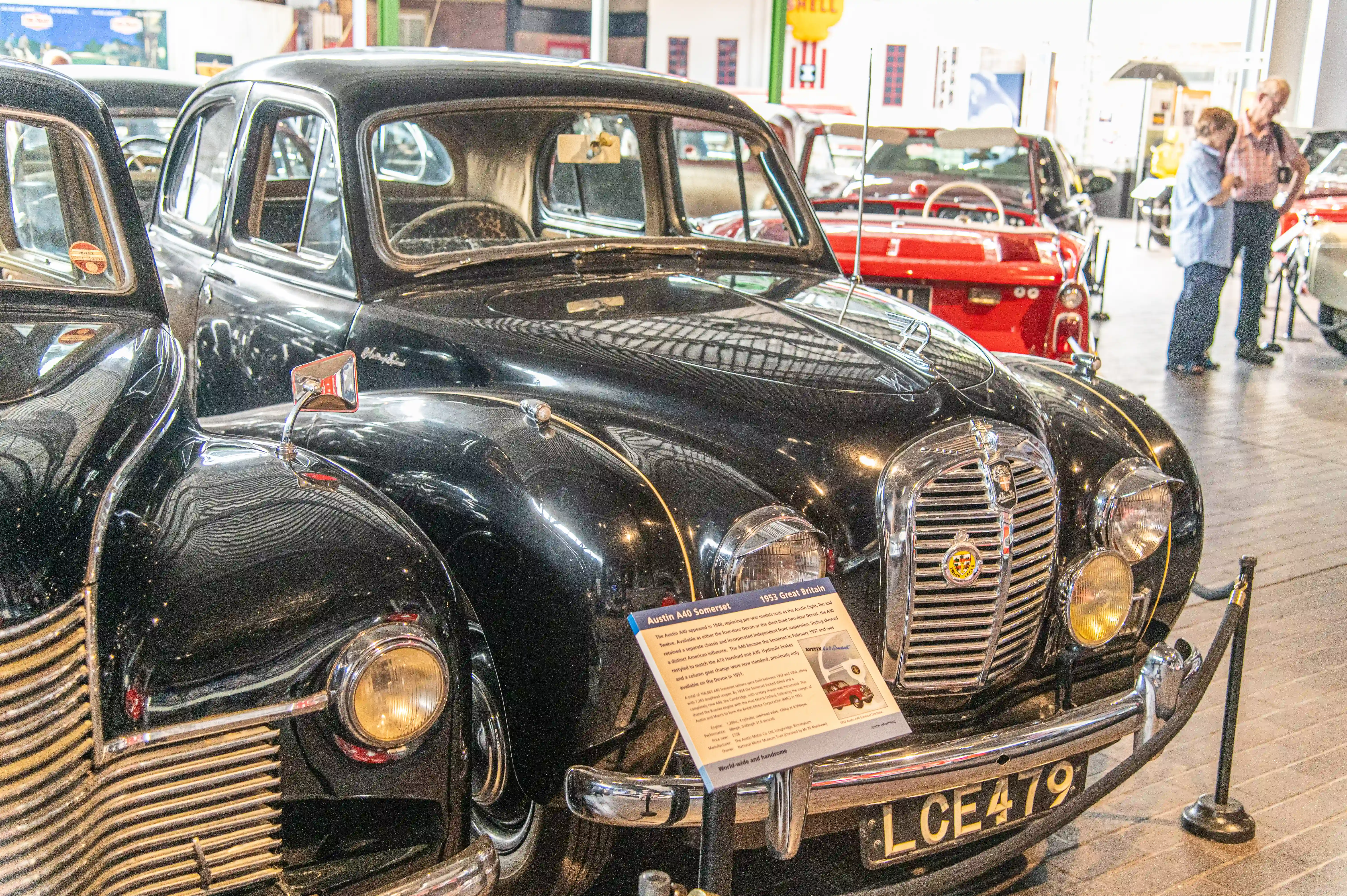The image shows a classic car, specifically a 1953 Austin A40 Somerset, displayed in a museum or showroom. The car is black with a distinctive grille and round headlights. There is an informational placard in front of the car providing details about its make and history. The setting appears to be an indoor exhibition with other vintage cars and people observing the displays.