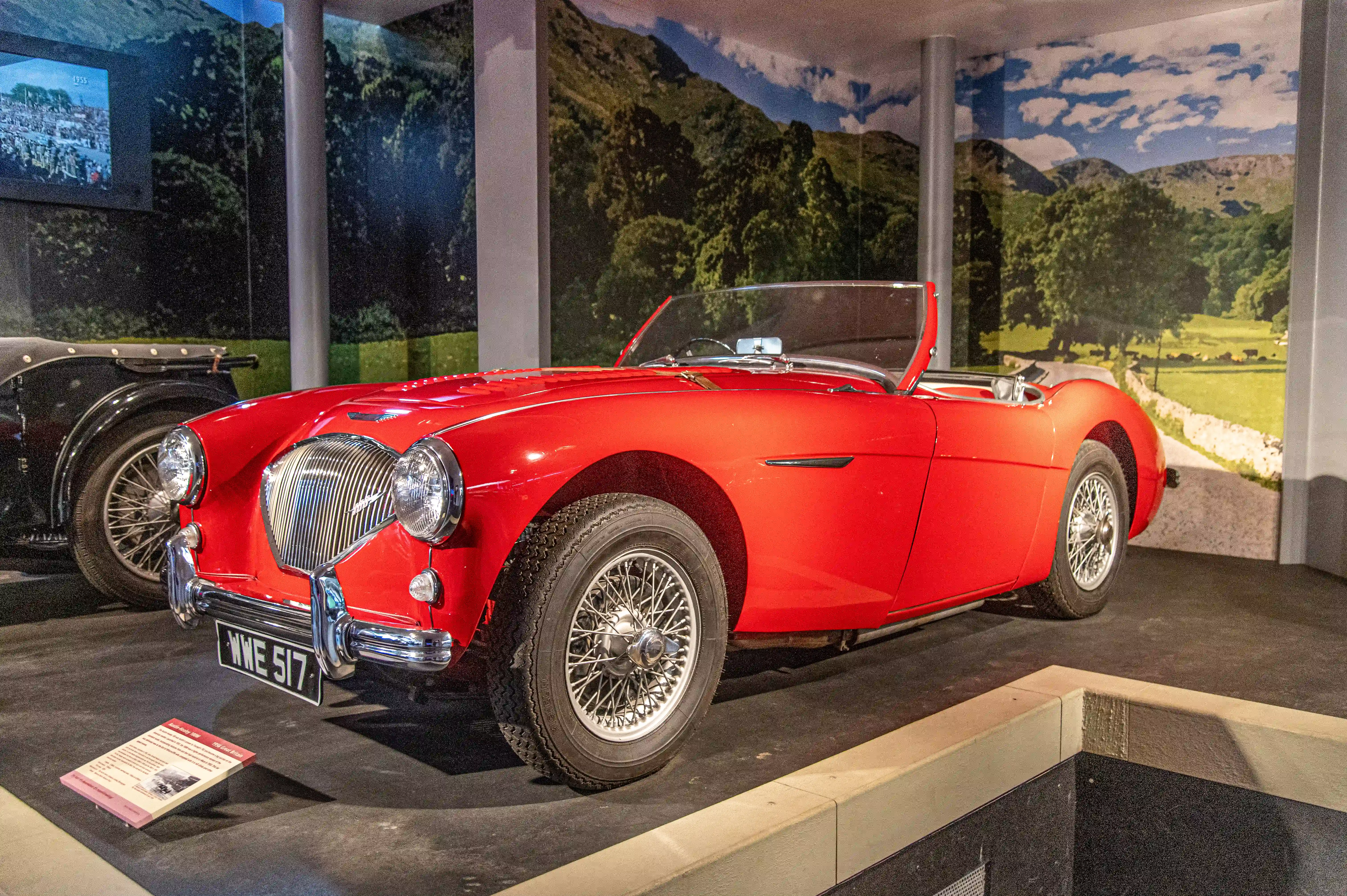 The image showcases a classic red convertible car displayed in a museum setting. The car has a vintage design with distinctive features such as wire-spoked wheels, dual headlights, and a chrome grille. The background features a scenic mural depicting a countryside landscape with trees, hills, and a partly cloudy sky. There is a small placard in front of the car providing information about it. Another vintage car is partially visible to the left of the red car.