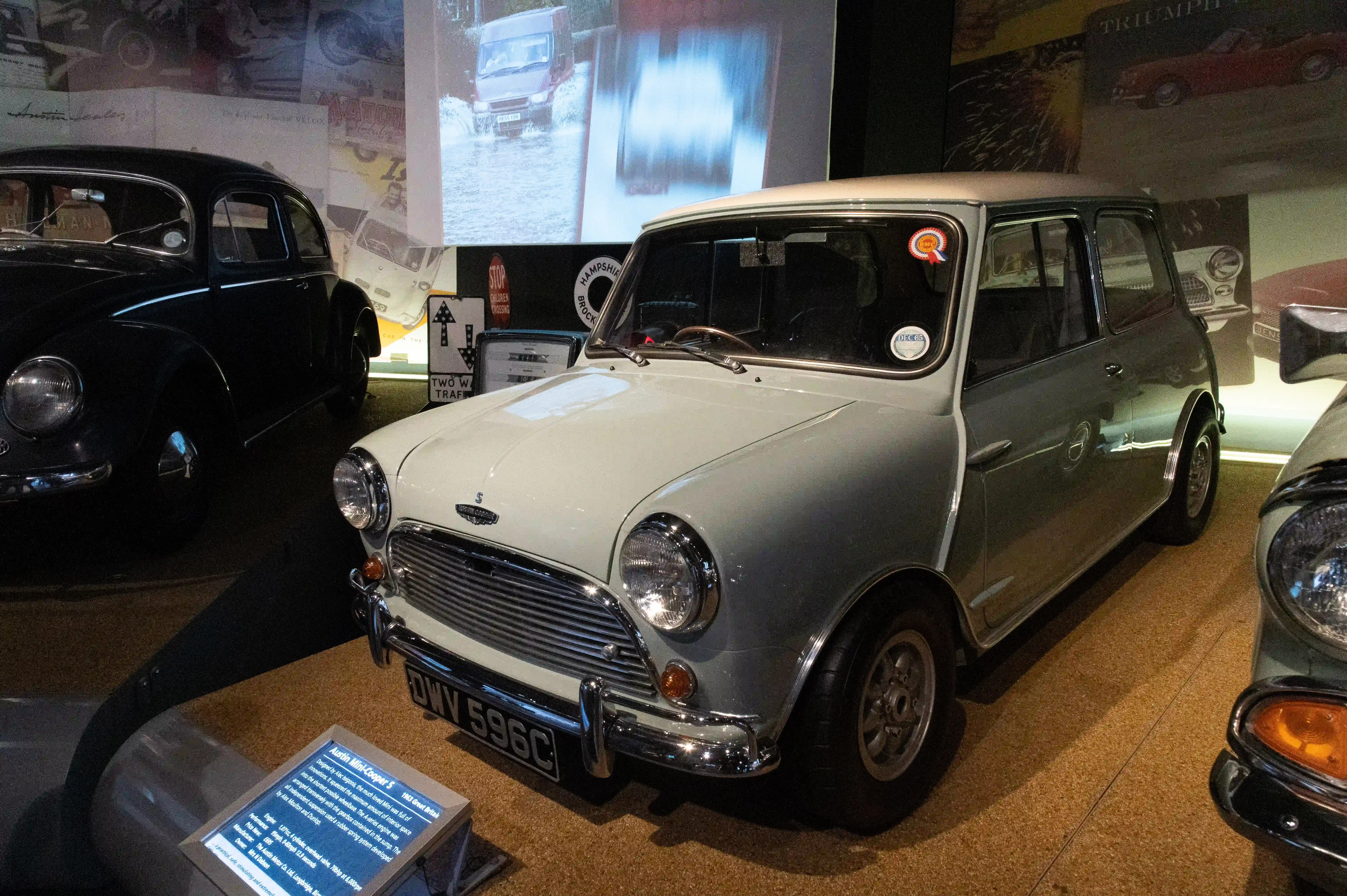 The image depicts a classic car exhibit, featuring a well-preserved Mini Cooper. The car is displayed in a museum-like setting with other vintage vehicles in the background. The Mini Cooper is a light grey color with distinctive round headlights and a chrome grille. There is an informational plaque in front of the car, providing details about its make and model. The setting includes various historical automotive advertisements and signs, enhancing the vintage atmosphere.