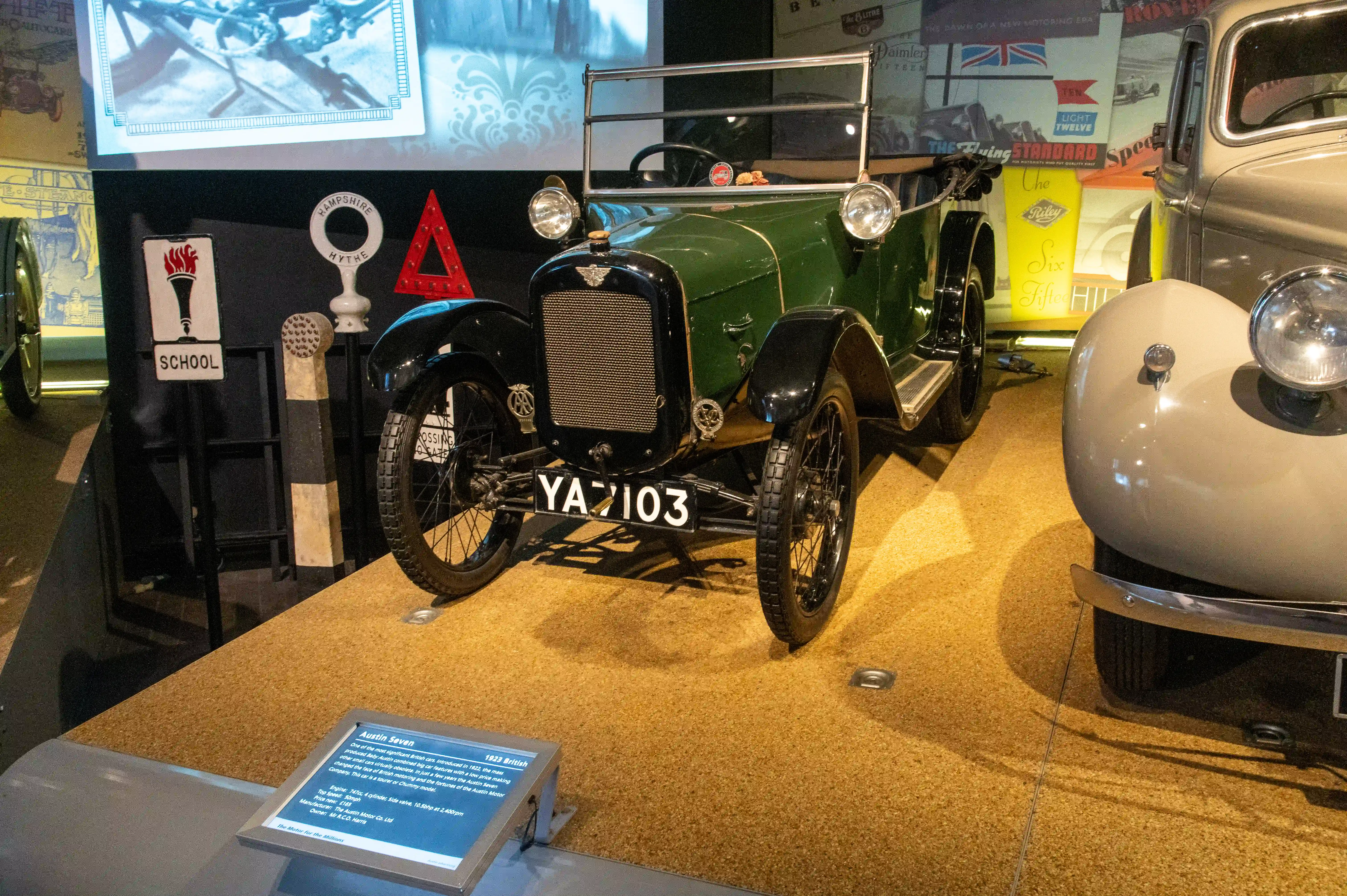 The image depicts a vintage car, specifically an Austin Seven from 1922, displayed in a museum. The car is green with black fenders and has the license plate 'YA 103'. An informational plaque in front of the car provides details about its historical significance and specifications. The display area includes other vintage vehicles and historical advertisements in the background.