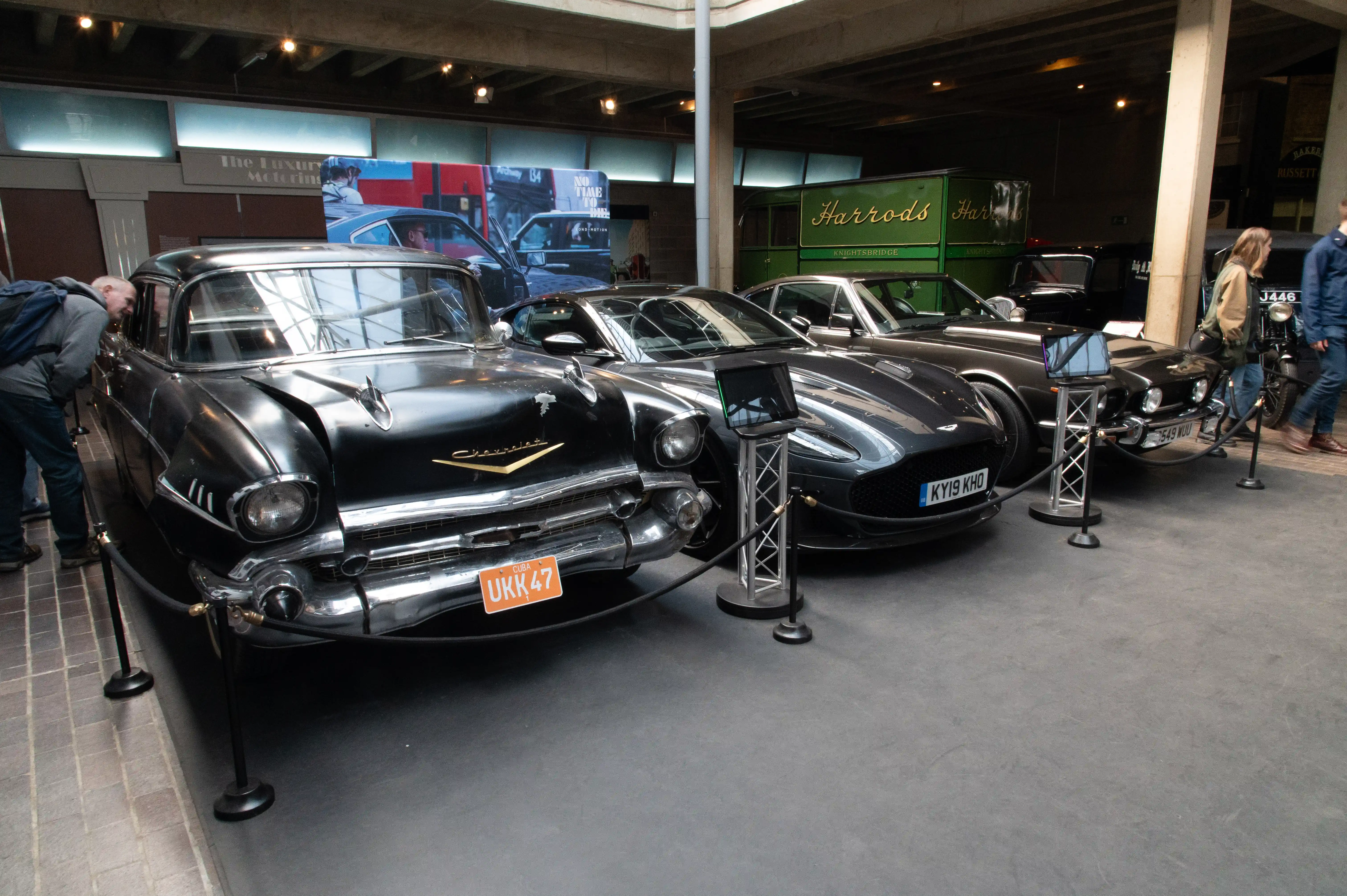 The image shows a classic car exhibition featuring vintage automobiles on display. The cars are cordoned off with stanchions and ropes to prevent close contact. Several people are observing and taking pictures of the cars. The setting appears to be an indoor showroom, with signs and banners in the background, including one from Harrods. The atmosphere suggests an event focused on luxury and classic automobiles.