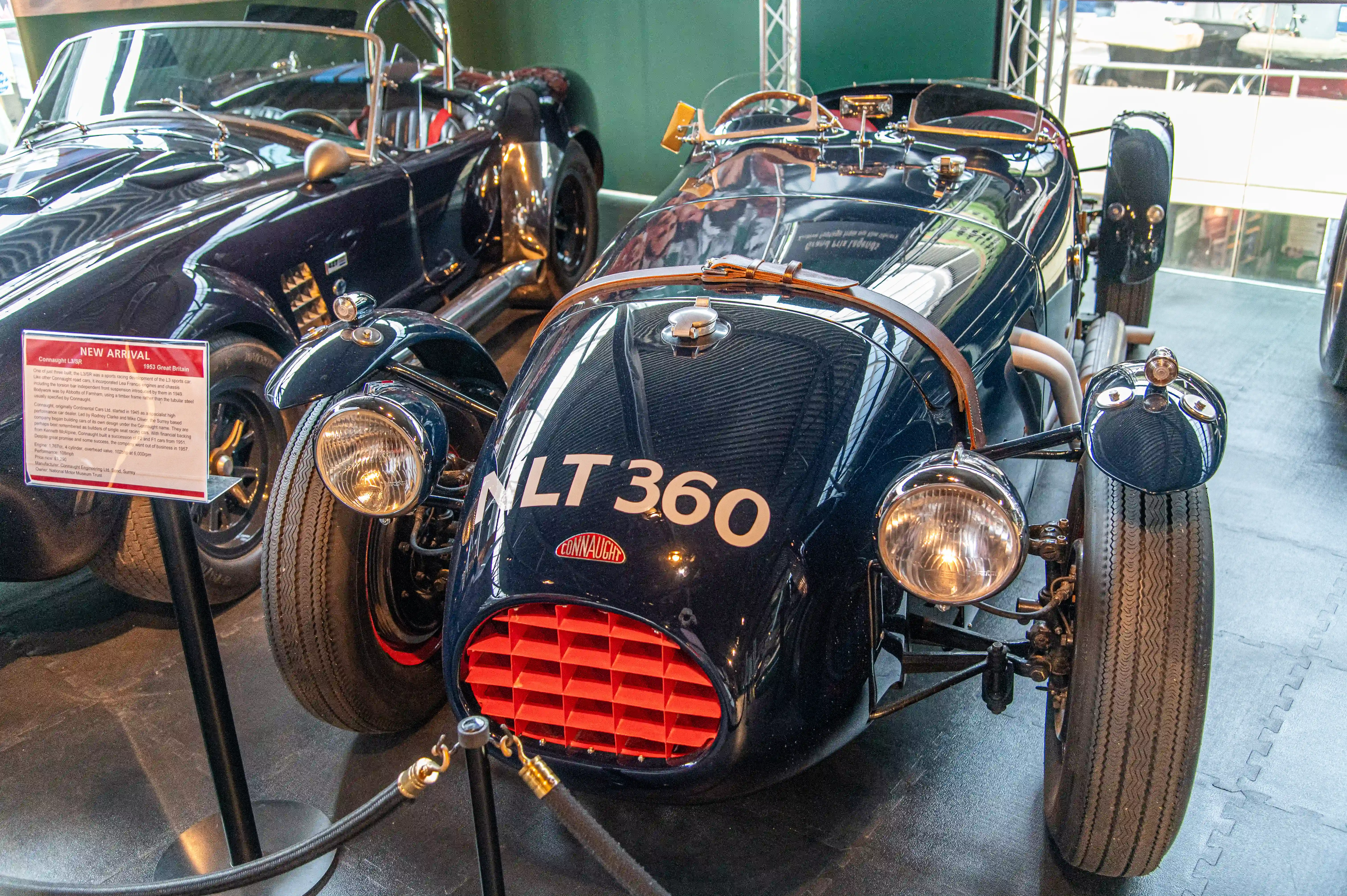 The image shows a classic car, specifically a 1933 SS100 Thundercloud Le Mans, on display in a museum. The car is black with the registration number ALT 360 and features a distinctive, streamlined design typical of vintage sports cars. There is an informational placard next to the car, indicating it is a new arrival and providing details about its history and specifications.