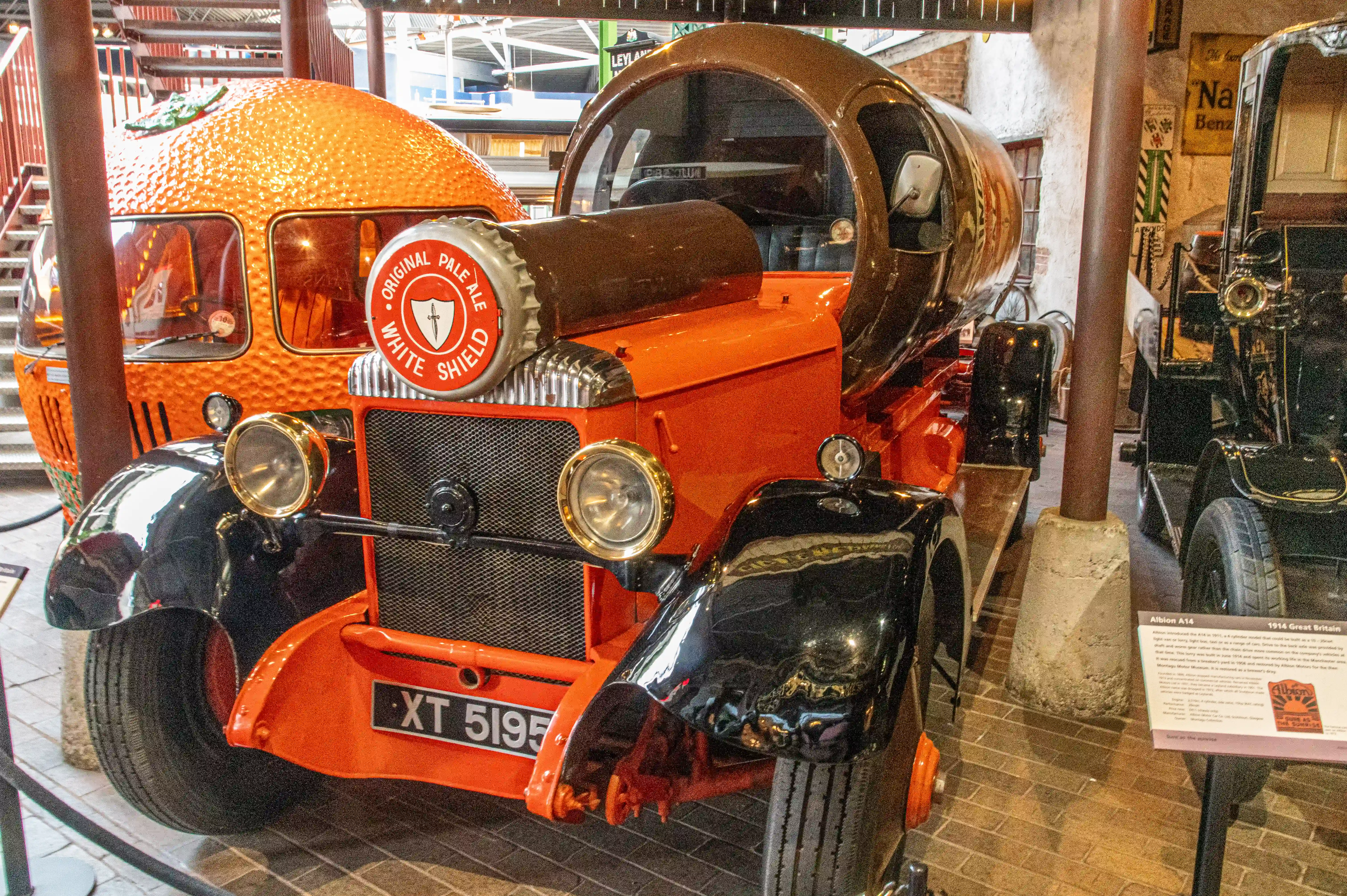 The image shows a vintage Albion A14 delivery truck from 1914, showcased in a museum. The truck is painted in a distinctive orange color with black fenders and features a large beer bottle-shaped hood, adorned with a label for 'Original Pale Ale White Shield'. The vehicle has a classic design with large headlights, a prominent grille, and a license plate reading 'XT 5195'. An informational plaque next to the truck provides details about its historical significance and specifications.