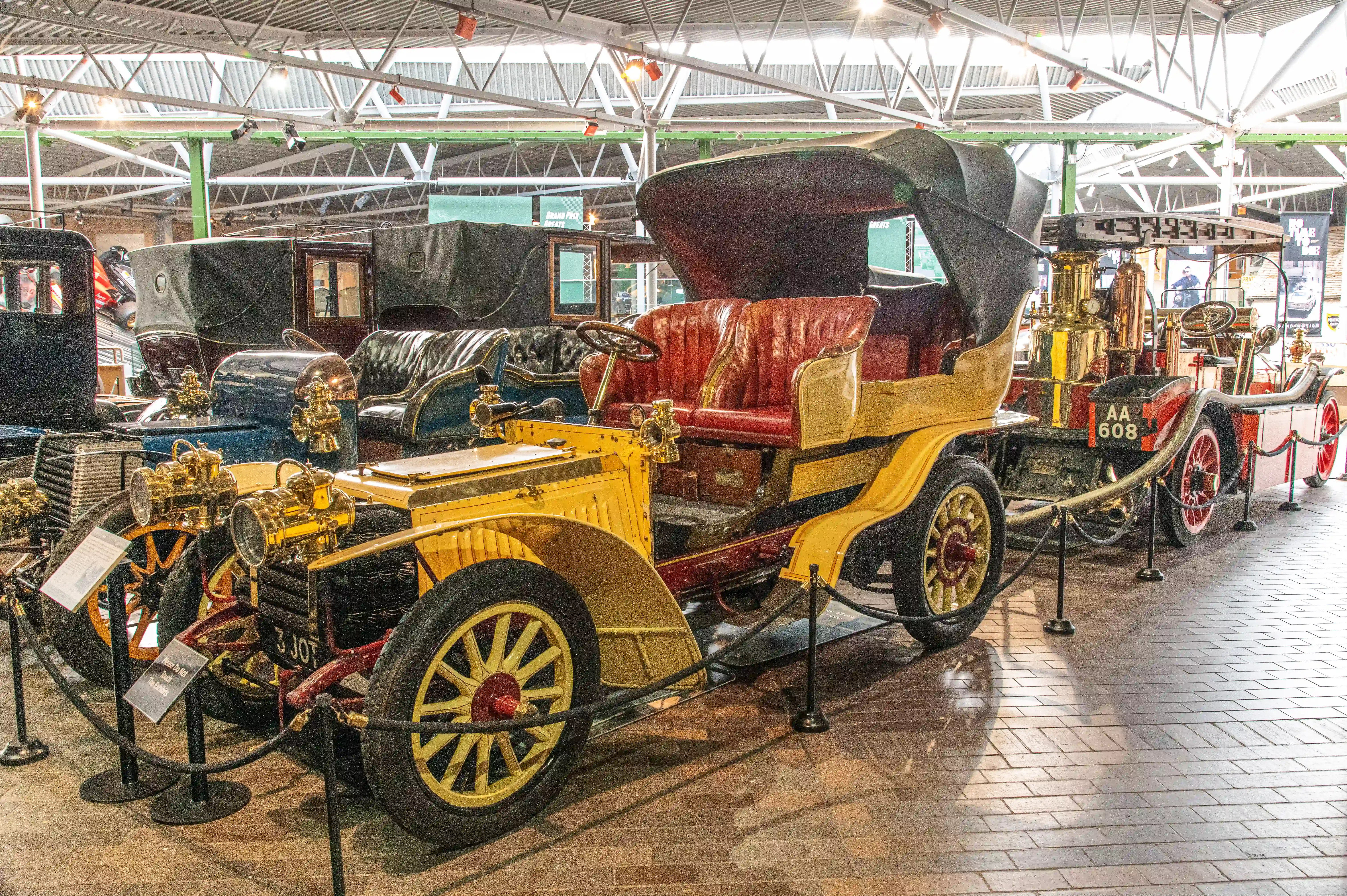 The image depicts a vintage car exhibition inside a spacious, well-lit hall. The central focus is on an antique yellow car with red leather seats, adorned with brass fixtures and large spoked wheels. The car is cordoned off with black ropes and stanchions, indicating it is part of a museum display. Other vintage vehicles are visible in the background, suggesting a collection of historical automobiles. Informational placards are placed near the cars, likely providing details about each exhibit.