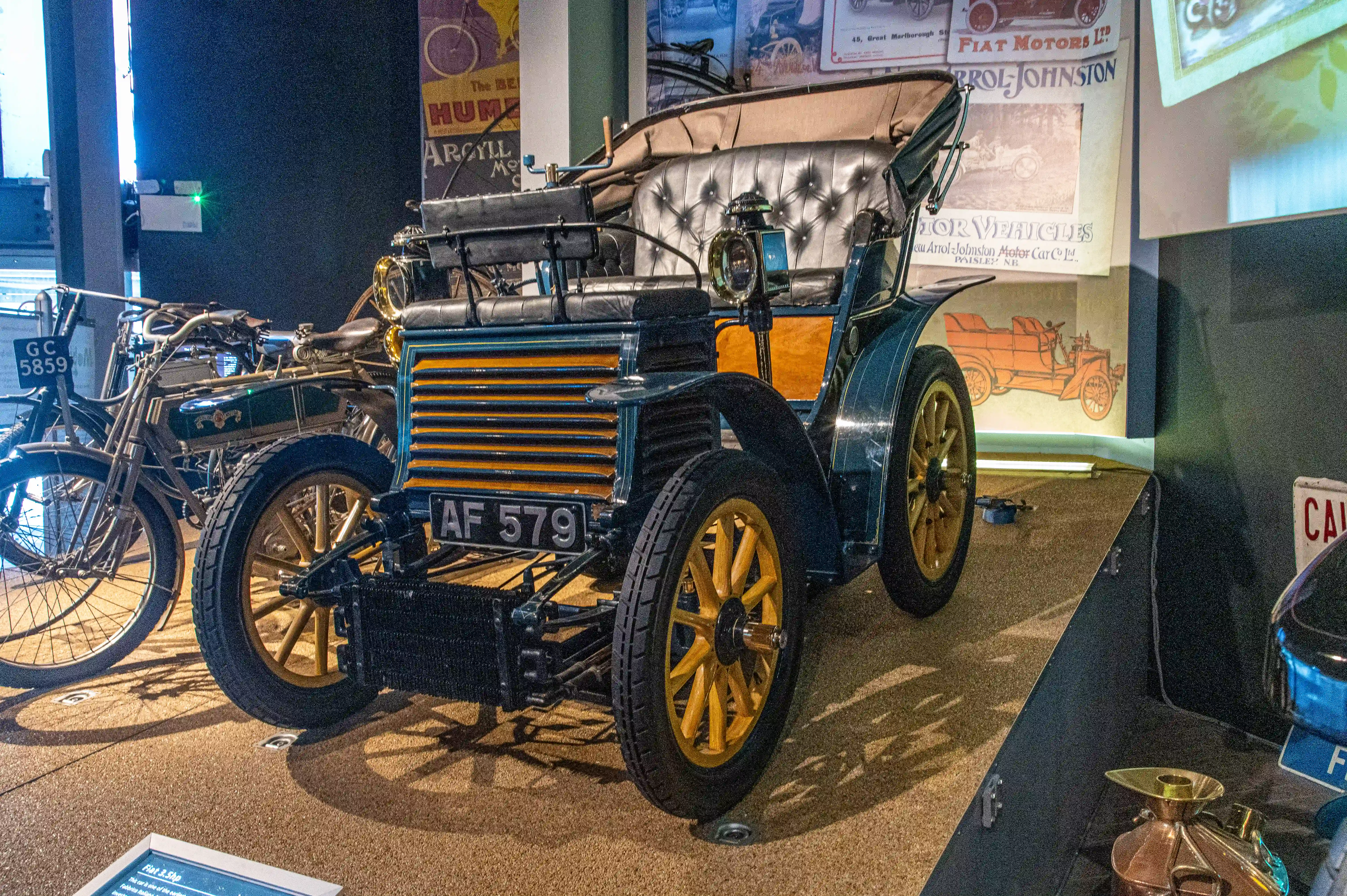 The image showcases a vintage car and a vintage bicycle on display in a museum. The car has a license plate reading 'AF 579' and features a classic design with large spoked wheels, a prominent front grille, and a tufted leather seat. The bicycle next to it has a license plate reading 'GC 5859' and a simpler, yet equally vintage design. The background includes various vintage advertisements and posters, enhancing the historical ambiance of the display.
