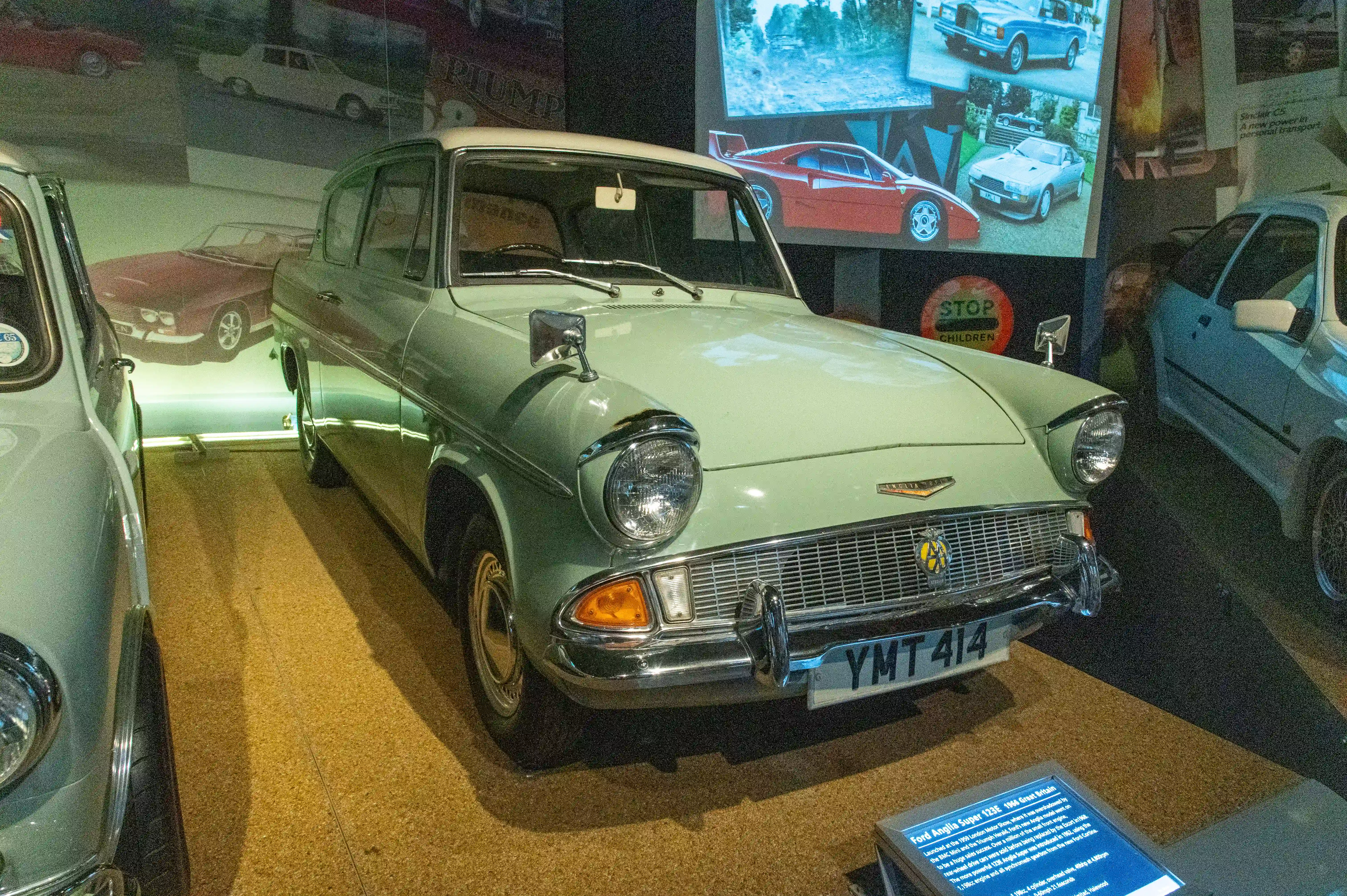 The image shows a classic car on display in a museum, specifically a Ford Anglia Super 123E from 1961. The car is painted in a light green color and is positioned centrally in the image. Surrounding the car are various informational displays and other vintage vehicles, contributing to the museum setting. The car's license plate reads 'YMT 414'.