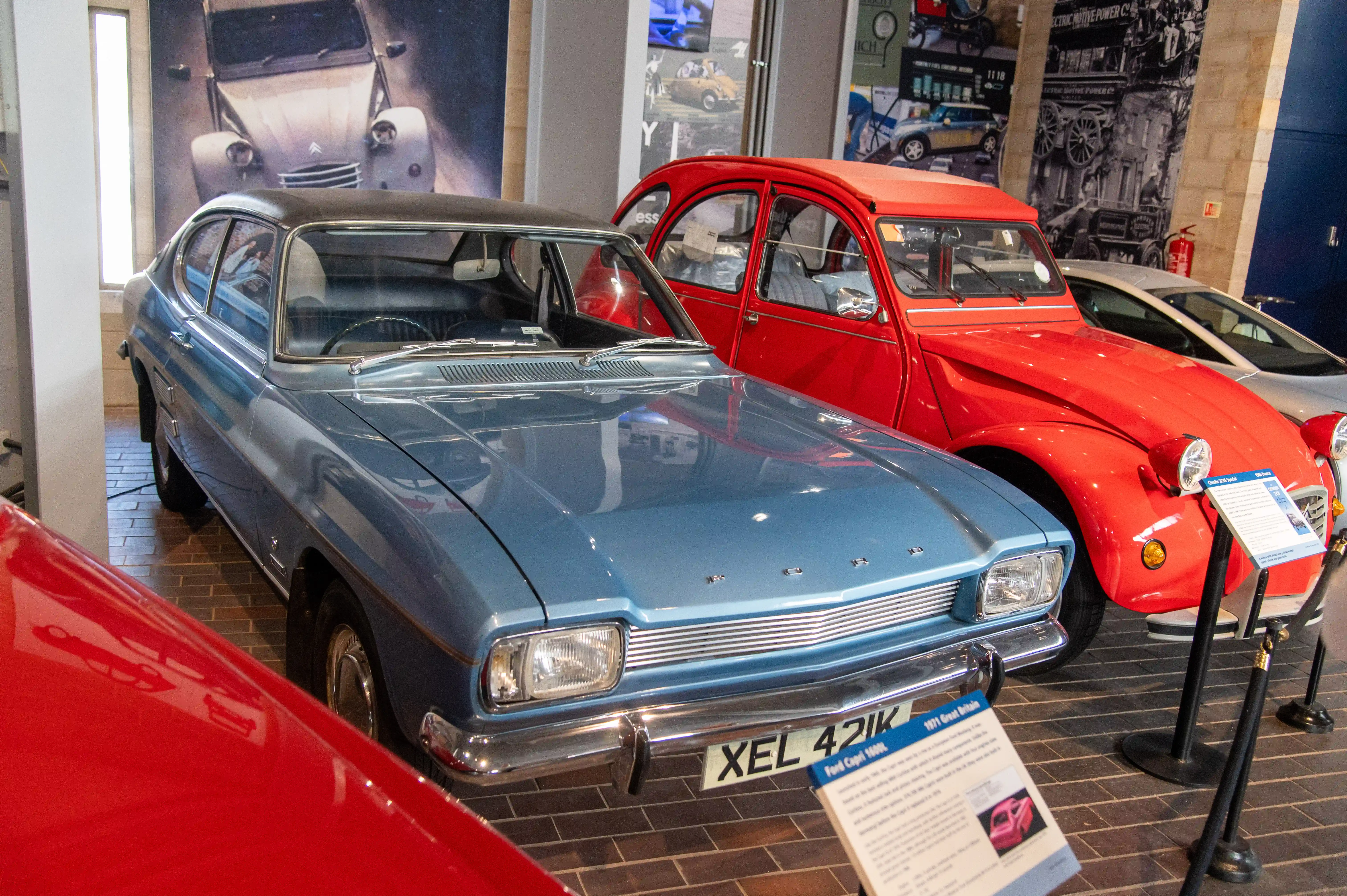 Here we have a couple of very distinctive cars! The star of the show is a rather sleek blue Ford Capri 1600L , with its chrome trim and a cool set of wheels. Next to it, stealing the scene with its vibrant colour, is a bright red Citroën 2CV. It's a really fun contrast between the two!
