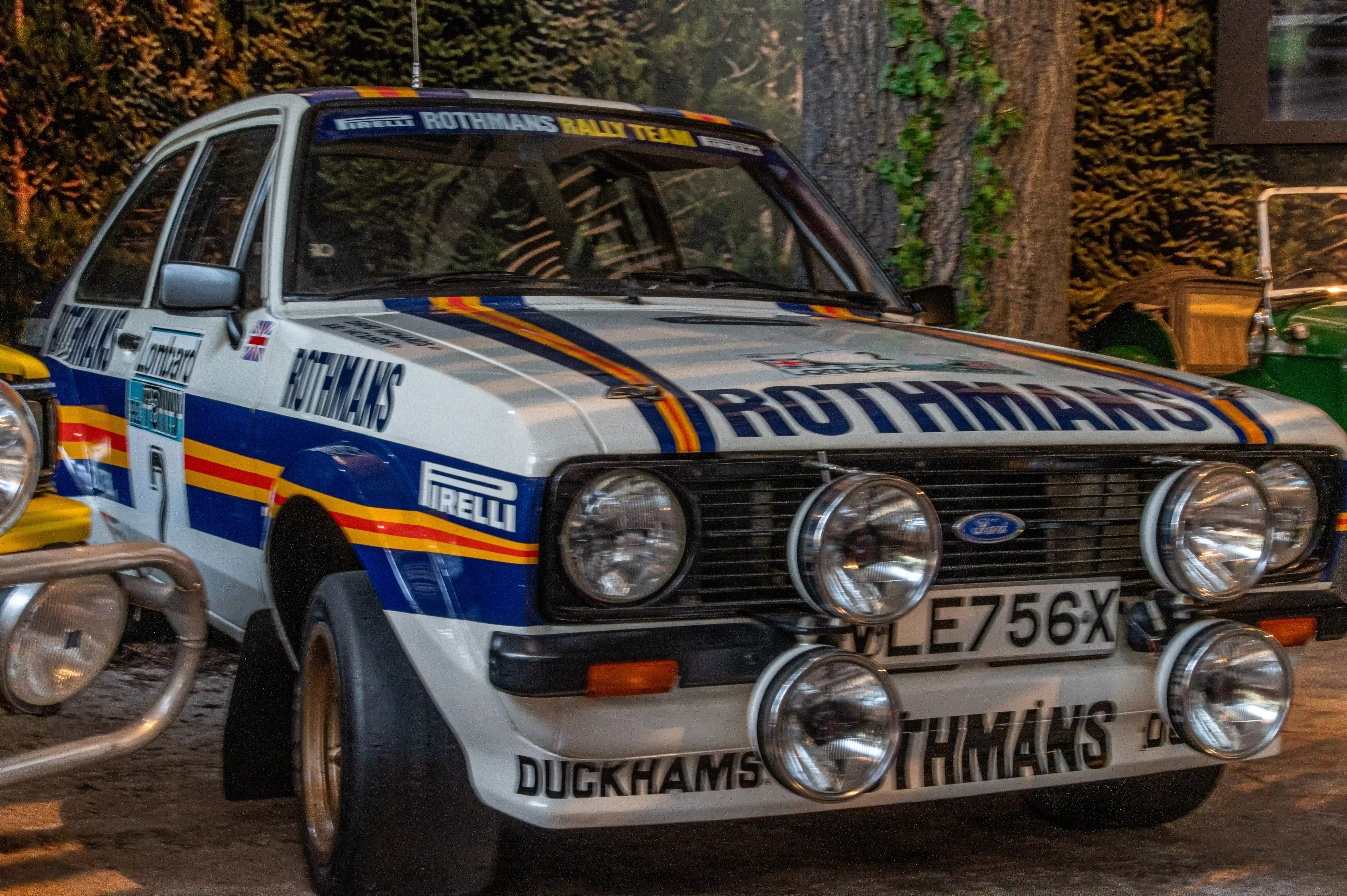 The image shows a classic rally car with the branding 'Rothmans Rally Team' prominently displayed on the hood and sides. The car features a white base color with blue, yellow, and red stripes. The vehicle has auxiliary lights mounted on the front bumper, and the license plate reads 'GVE756X'. The car appears to be a Ford, as indicated by the emblem on the grille. The overall design and decals suggest it is a vintage rally car, likely from a historical motorsport event.