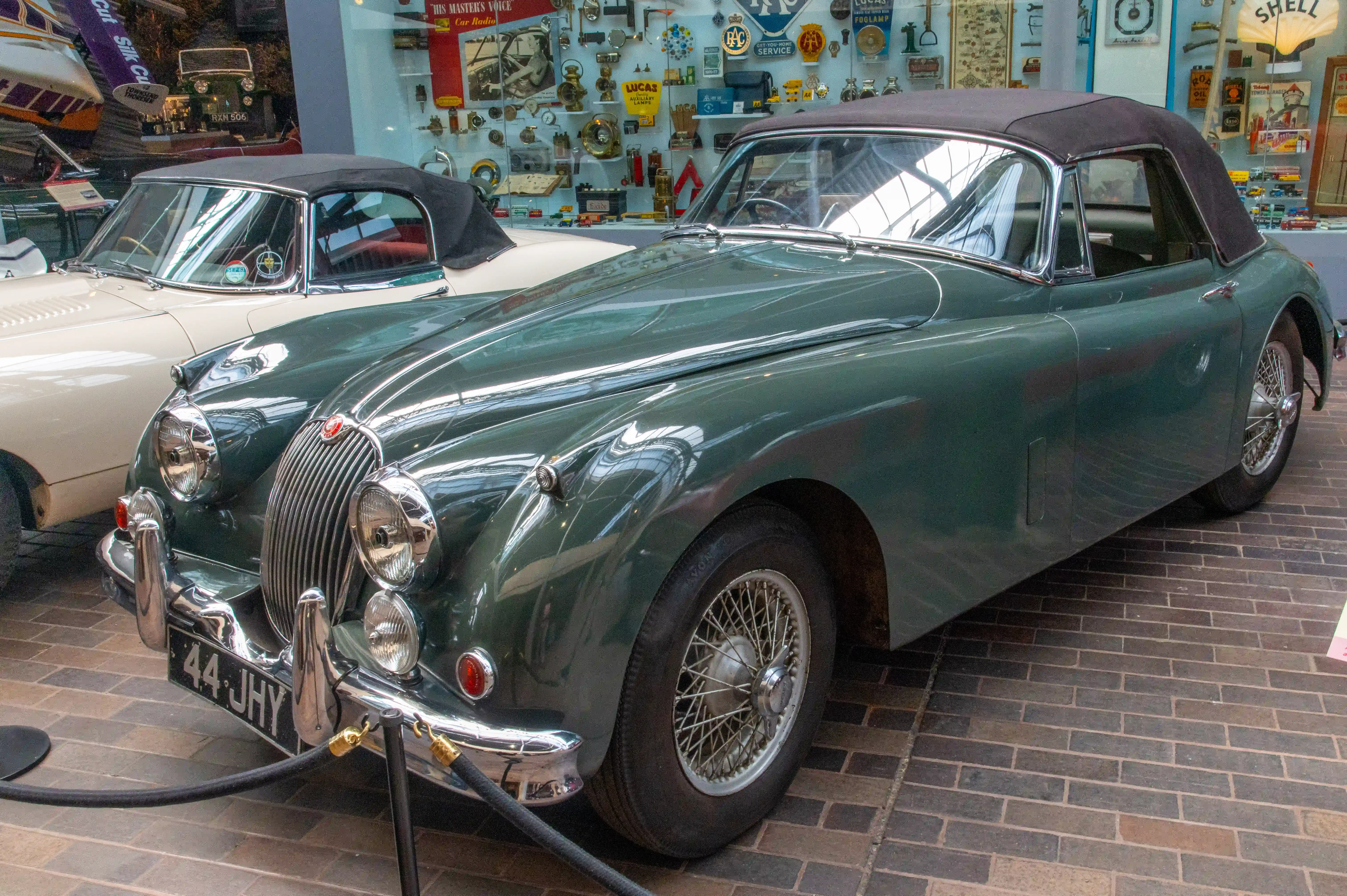 The car is dark green with a black convertible top and wire-spoke wheels. It is parked on a brick floor with another classic car, a cream-colored vehicle, visible in the background. The background features various automotive memorabilia, including signs, tools, and other vintage car parts. The setting suggests a celebration of automotive history and classic car enthusiasts.