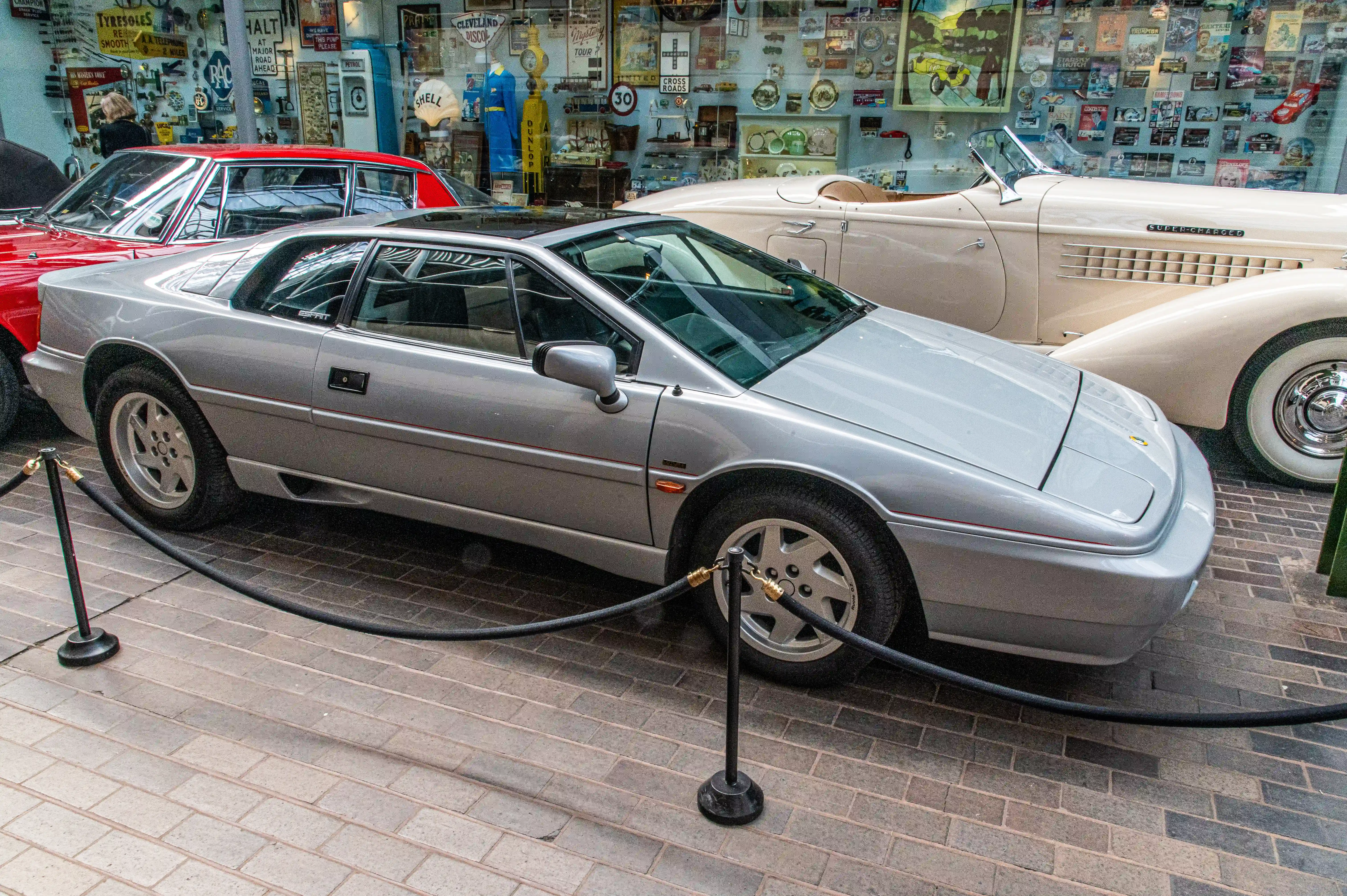 The image shows a silver sports car displayed in a museum or exhibition setting. The car is cordoned off with velvet ropes and stanchions, indicating it is part of a collection. The background features a wall filled with various automotive memorabilia, including vintage advertisements, signs, and posters. There are other classic cars visible in the background, suggesting a theme of automotive history or classic cars. The setting appears to be indoors, with a brick floor and a glass wall showcasing the items.