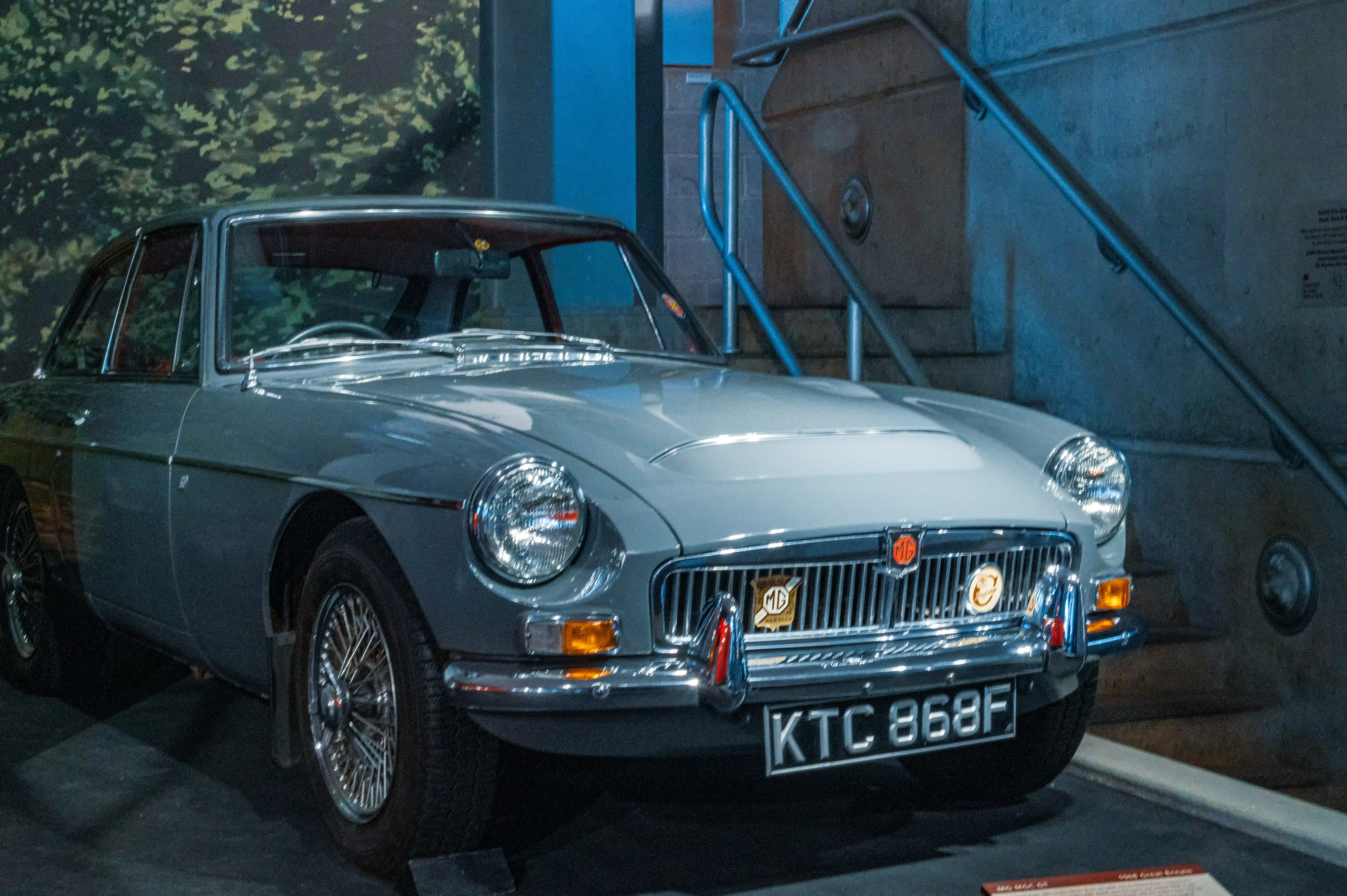 The image shows a classic car, specifically a light blue MG sports car, on display in a museum. The car has a convertible top, wire-spoke wheels, and a distinctive chrome grille with vertical slats. The license plate reads 'KTC 868F,' and there is a badge on the front of the car that reads 'MD.' The car is positioned in front of a blue railing and a large window, with another vehicle partially visible in the background.