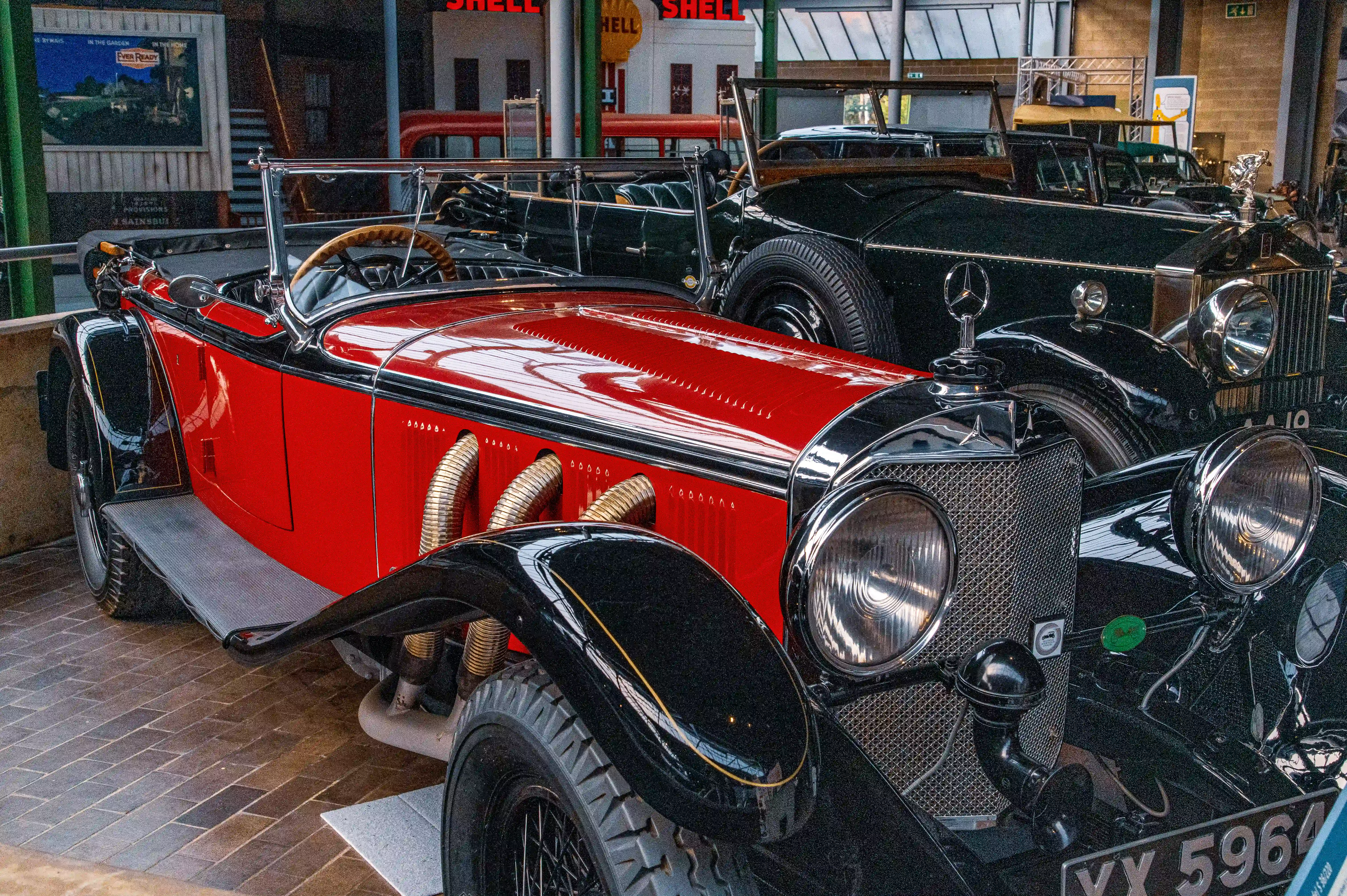 The image showcases a vintage car with a red body and black fenders, featuring prominent headlights and a distinctive grille. The car is displayed in a museum-like setting with other classic vehicles in the background. The setting appears to be an old-fashioned gas station, complete with Shell signage and various vintage advertisements. The car's license plate reads 'VY 596'.