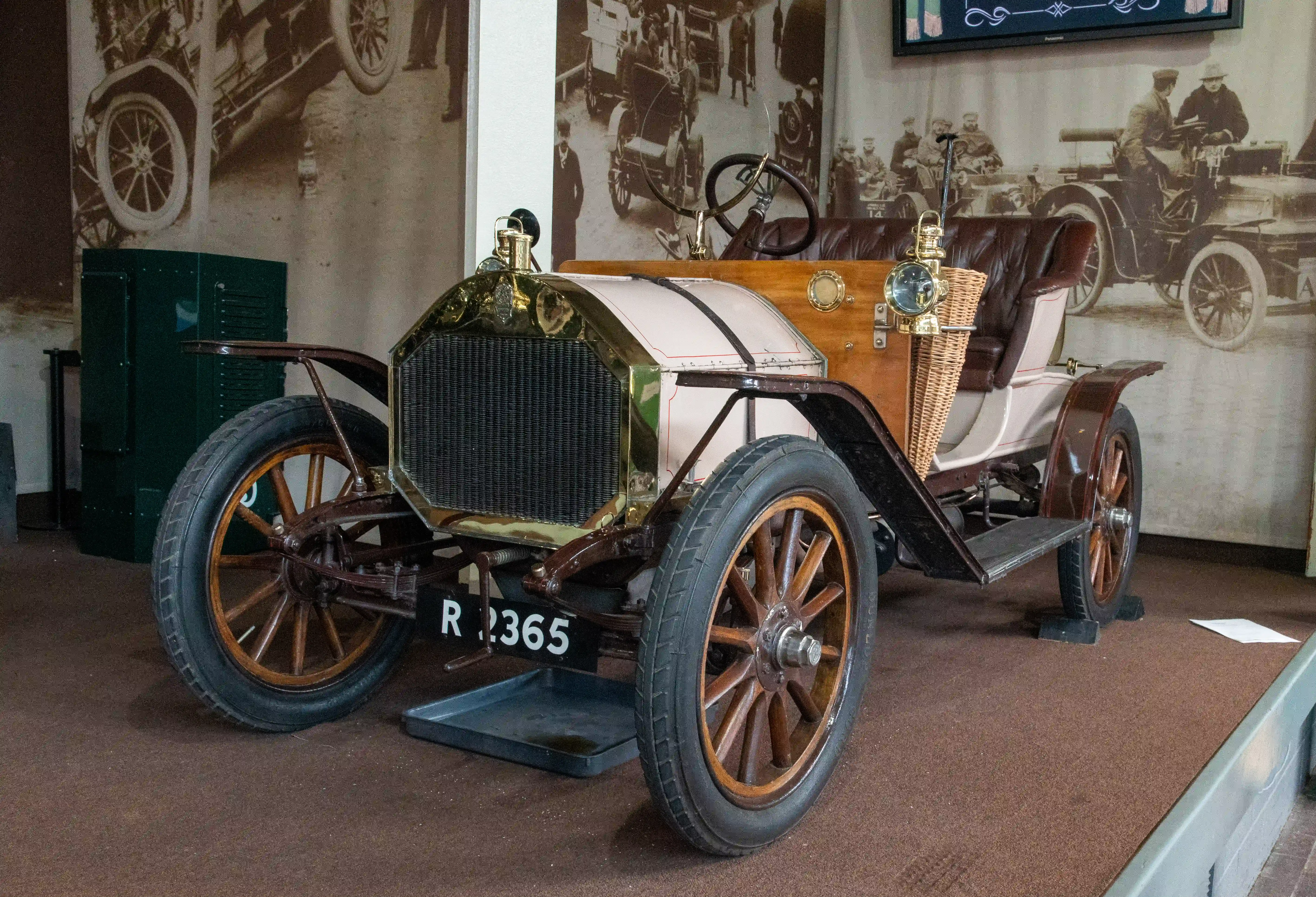 The image depicts an antique car on display in a museum. The car has a vintage design with large spoked wheels, a wooden frame, and a simple engine compartment. The vehicle features early automotive technology, including a large front grille and minimal bodywork. The background includes black-and-white photographs of early automobiles and their development. The car is positioned on a raised platform with a license plate reading 'R 2365'.