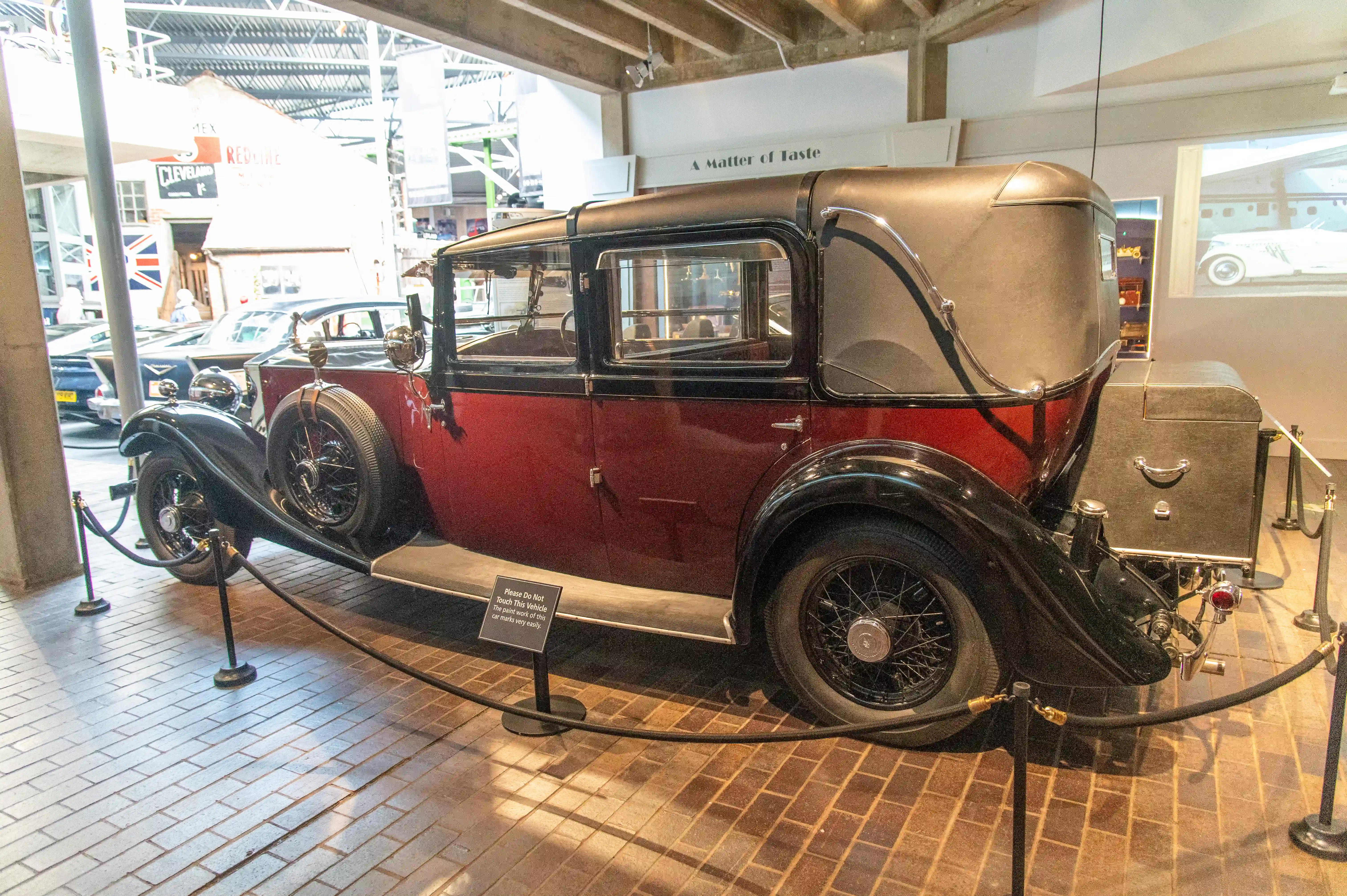 The image depicts a classic car displayed in a museum. The car is a maroon and black two-door sedan with a vintage design, featuring wire-spoked wheels and large fenders. It is cordoned off with velvet ropes and has a sign requesting visitors not to touch. The setting appears to be an indoor exhibit with other vintage vehicles and memorabilia in the background.