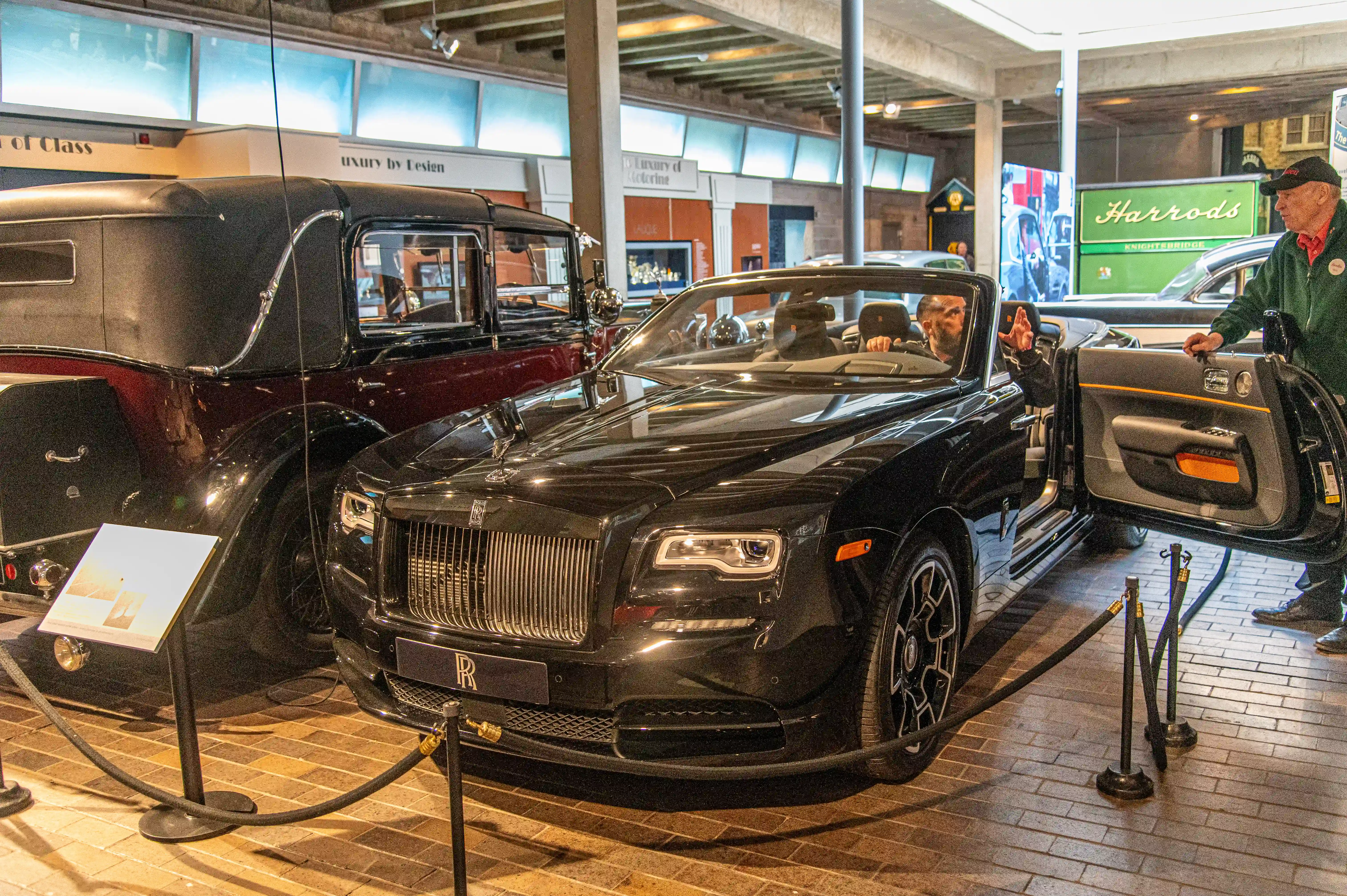The image depicts a luxury car exhibition featuring a prominent black Rolls-Royce with its door open, showcasing its interior. The car is displayed in an indoor setting, likely a showroom or museum, with informational placards nearby. In the background, there are other vintage and luxury cars, and a sign for Harrods, indicating a high-end, sophisticated atmosphere. Two individuals are present, one seated inside the Rolls-Royce and another standing by the open door, engaging with the exhibit.