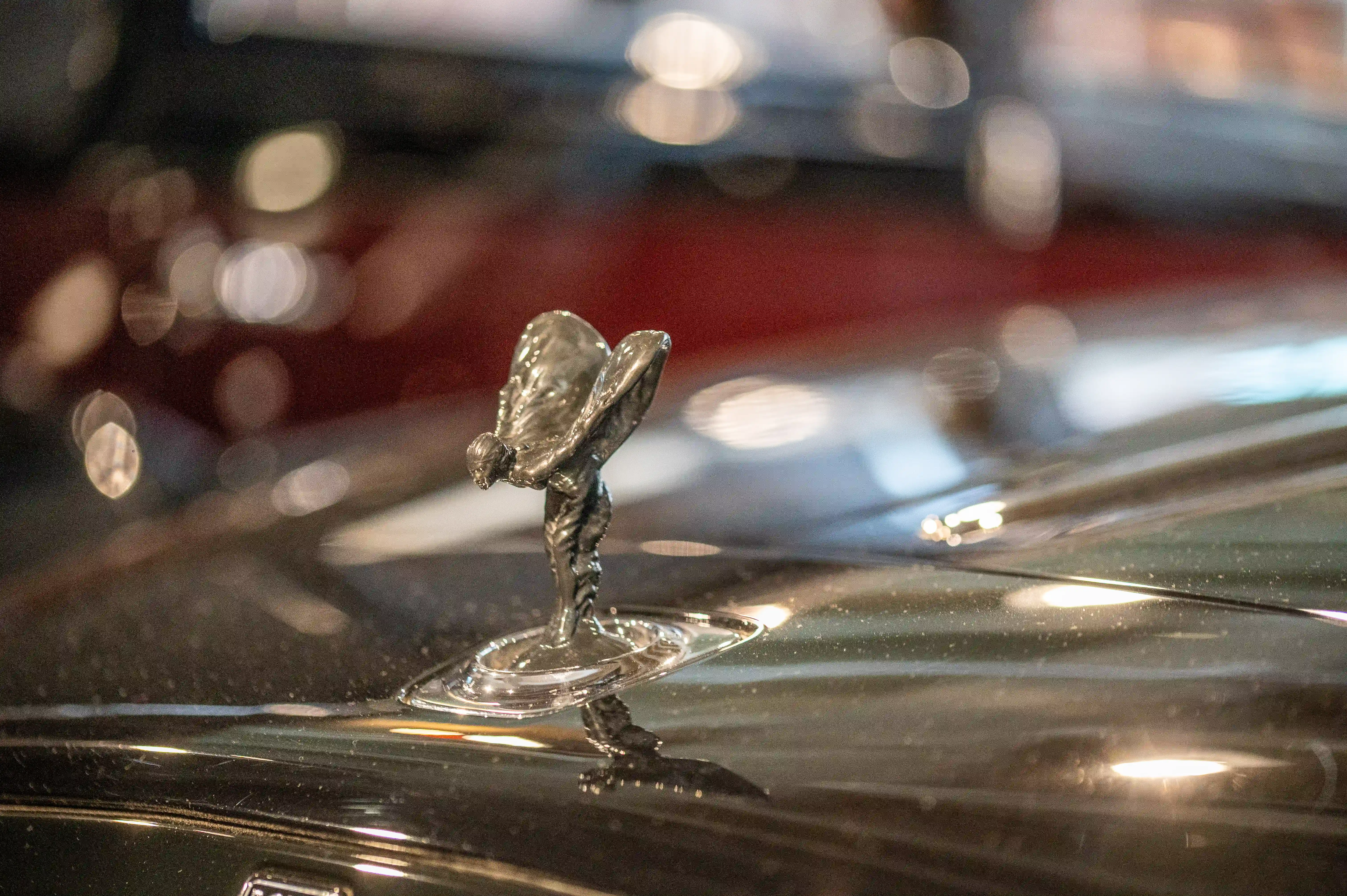 The image shows a close-up of the iconic flying lady mascot, also known as the Spirit of Ecstasy, on the hood of a Rolls-Royce car. The mascot is a symbol of the luxury and elegance associated with the Rolls-Royce brand. The background is blurred, emphasizing the intricate details of the mascot. The car's hood is slightly wet, suggesting it might have been raining or the car has been recently washed. The overall scene conveys a sense of sophistication and high-end craftsmanship.