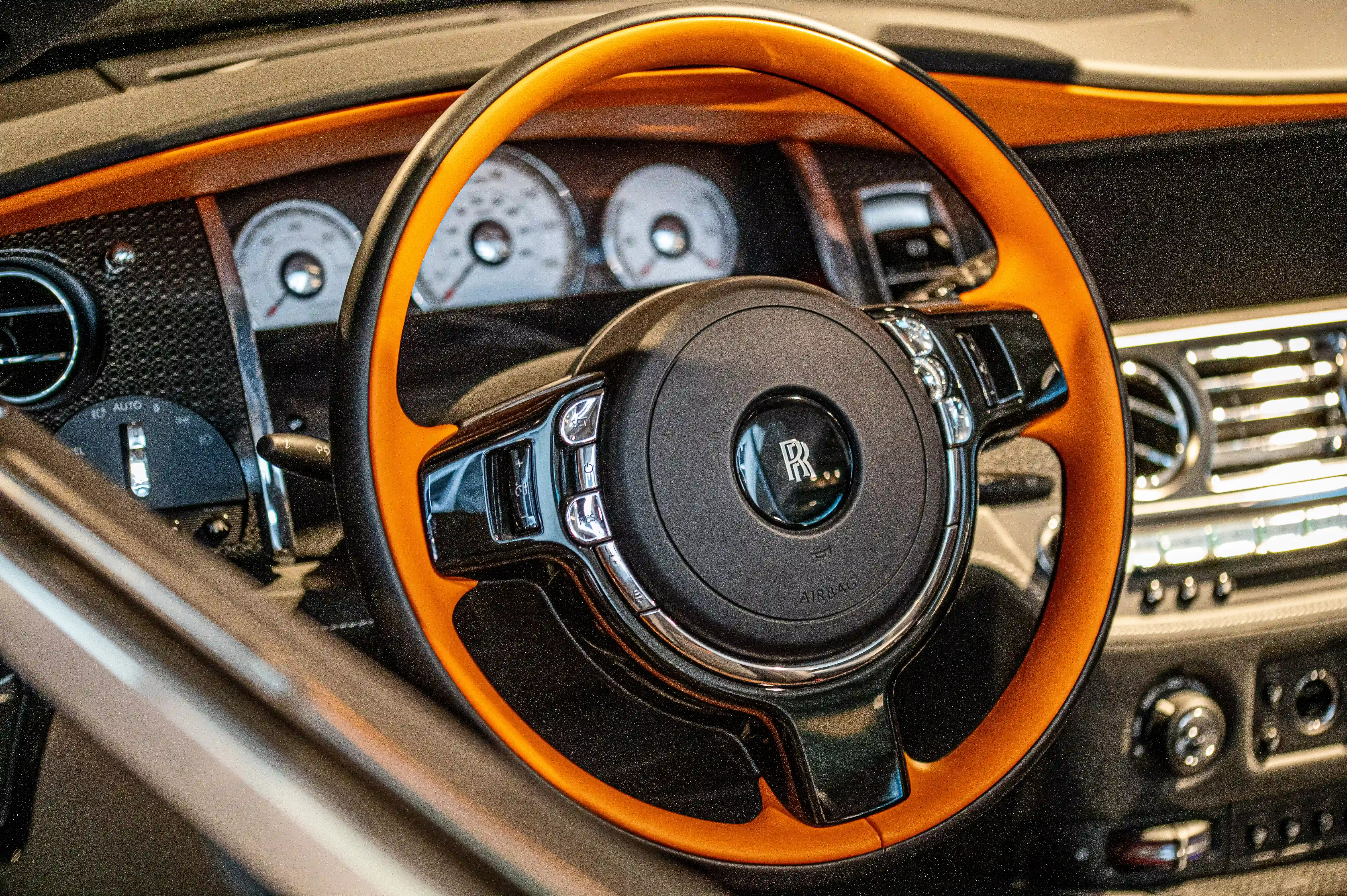 The image shows the interior of a luxury car, specifically focusing on the steering wheel and dashboard. The steering wheel is wrapped in a combination of black leather and has an orange accent stitching. The dashboard features several gauges and dials, including a tachometer and speedometer, and the car's branding is prominently displayed in the center of the steering wheel. The overall design and materials suggest a high-end, possibly classic or vintage vehicle.
