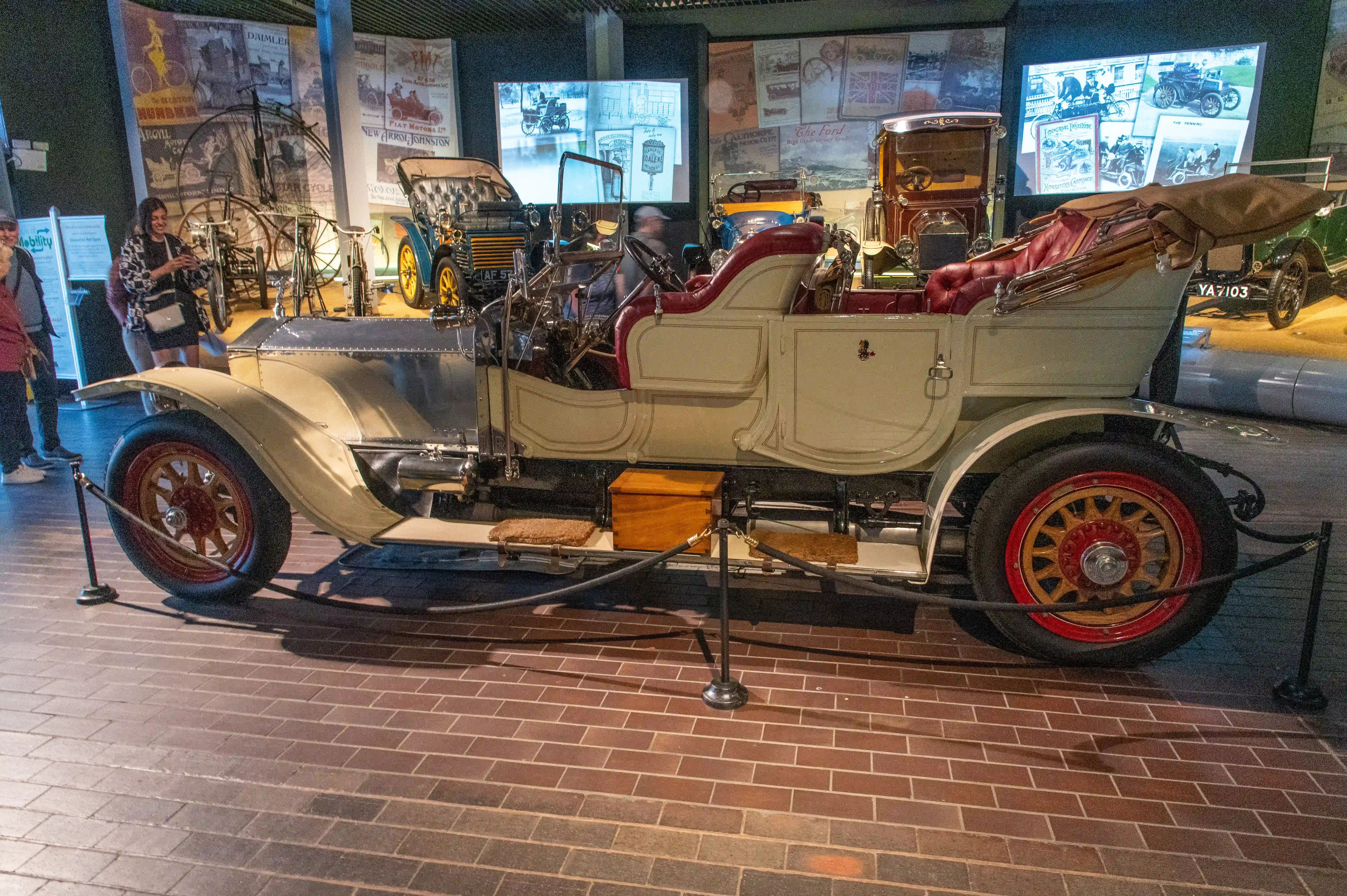 The image depicts a vintage car on display in a museum. The car features a beige body with red upholstery and a convertible top. It has large, spoked wheels with red rims and is surrounded by a rope barrier to prevent visitors from touching it. In the background, there are various posters and advertisements related to early automotive history. A few people are seen observing the exhibits, and other vintage vehicles are visible in the display area.