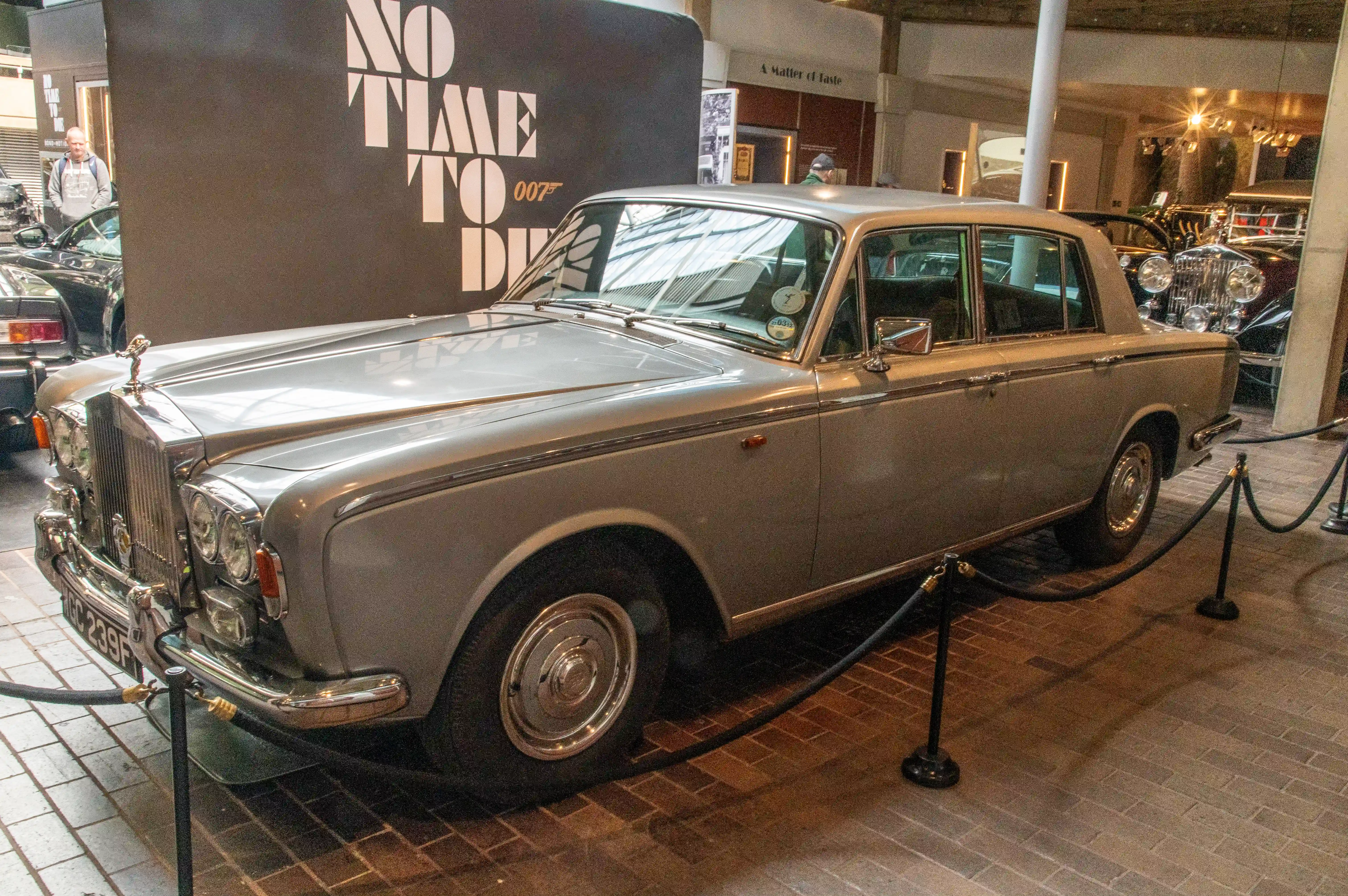 The image shows a classic silver Rolls-Royce car on display in a museum, cordoned off with stanchions and ropes. The car has a distinctive front grille with the Spirit of Ecstasy hood ornament and round headlights. Behind the car, there is a large sign with the text 'NO TIME TO DIE' and '007', indicating a connection to the James Bond film series. The car's license plate reads '3367 F'. The setting appears to be an exhibition or a museum dedicated to James Bond, featuring various vehicles and artifacts from the films.