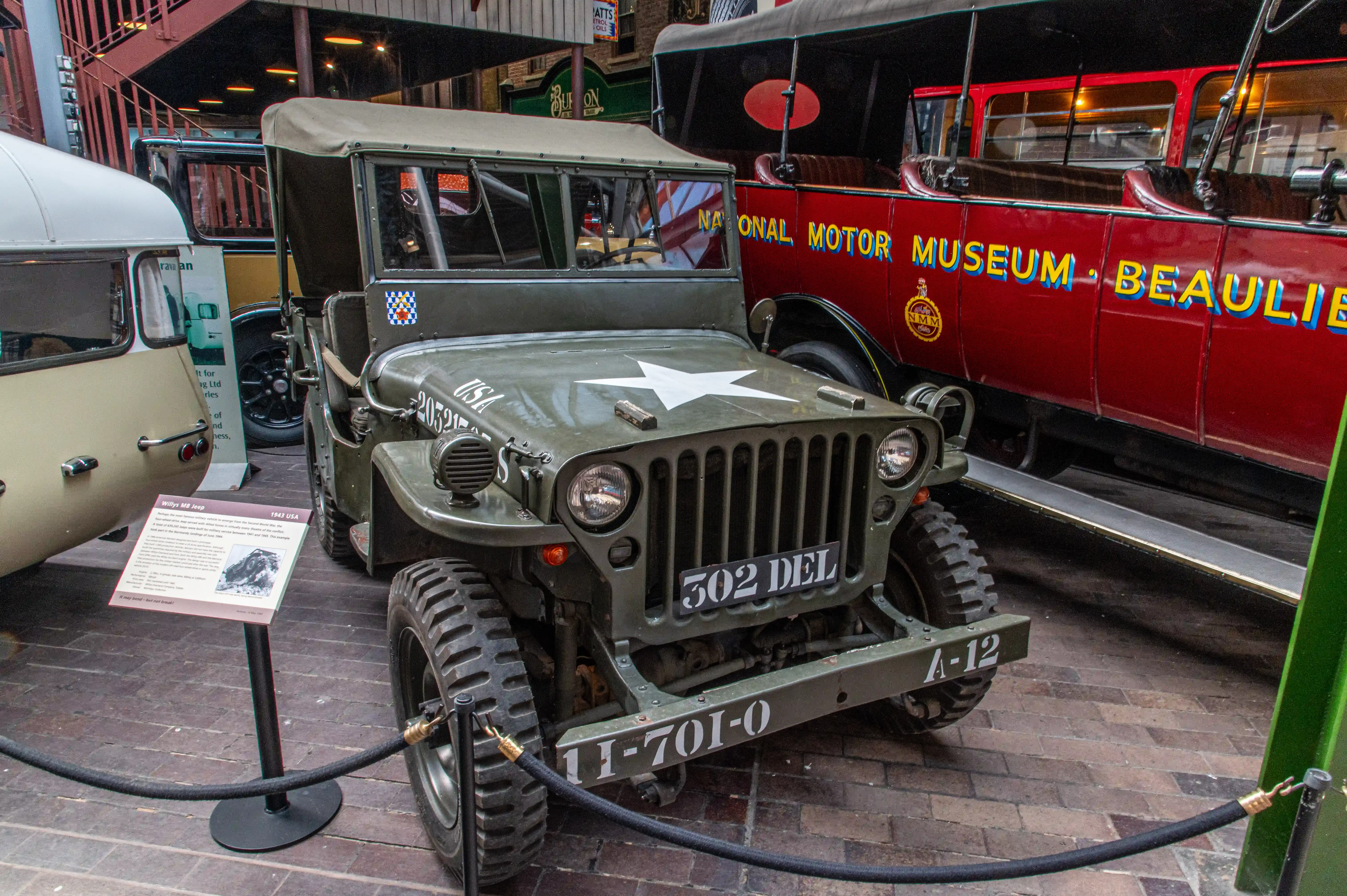 The image shows a vintage military jeep on display at the National Motor Museum in Beaulieu. The jeep, identified as a 1943 USA model, features a green body with a white star on the hood, and various markings including '502 DEL' on the front and 'II-701-0' on the bumper. It is cordoned off with a rope barrier, and an informational placard is positioned in front of it. Other vehicles, including a red bus and a beige van, are also visible in the background.