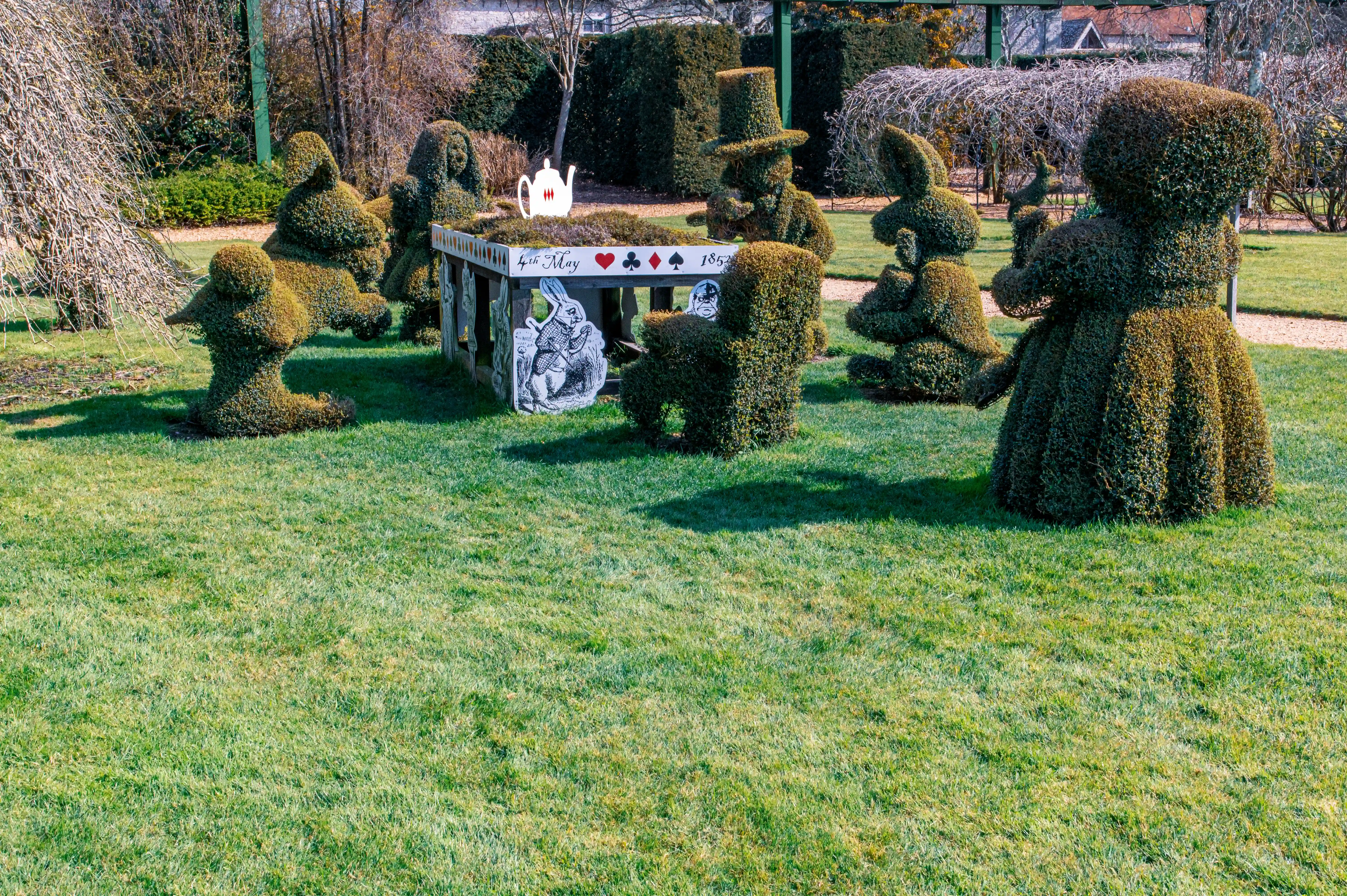The image depicts a topiary garden scene inspired by 'Alice in Wonderland.' It features hedges trimmed into the shapes of characters from the story, including Alice and the Mad Hatter, seated around a table. The table is adorned with playing cards and a teapot, and there is a sign indicating '4th May 1862,' the date of the Mad Hatter's tea party in the book. The scene is set in a well-maintained garden with green grass and various other plants and hedges in the background.