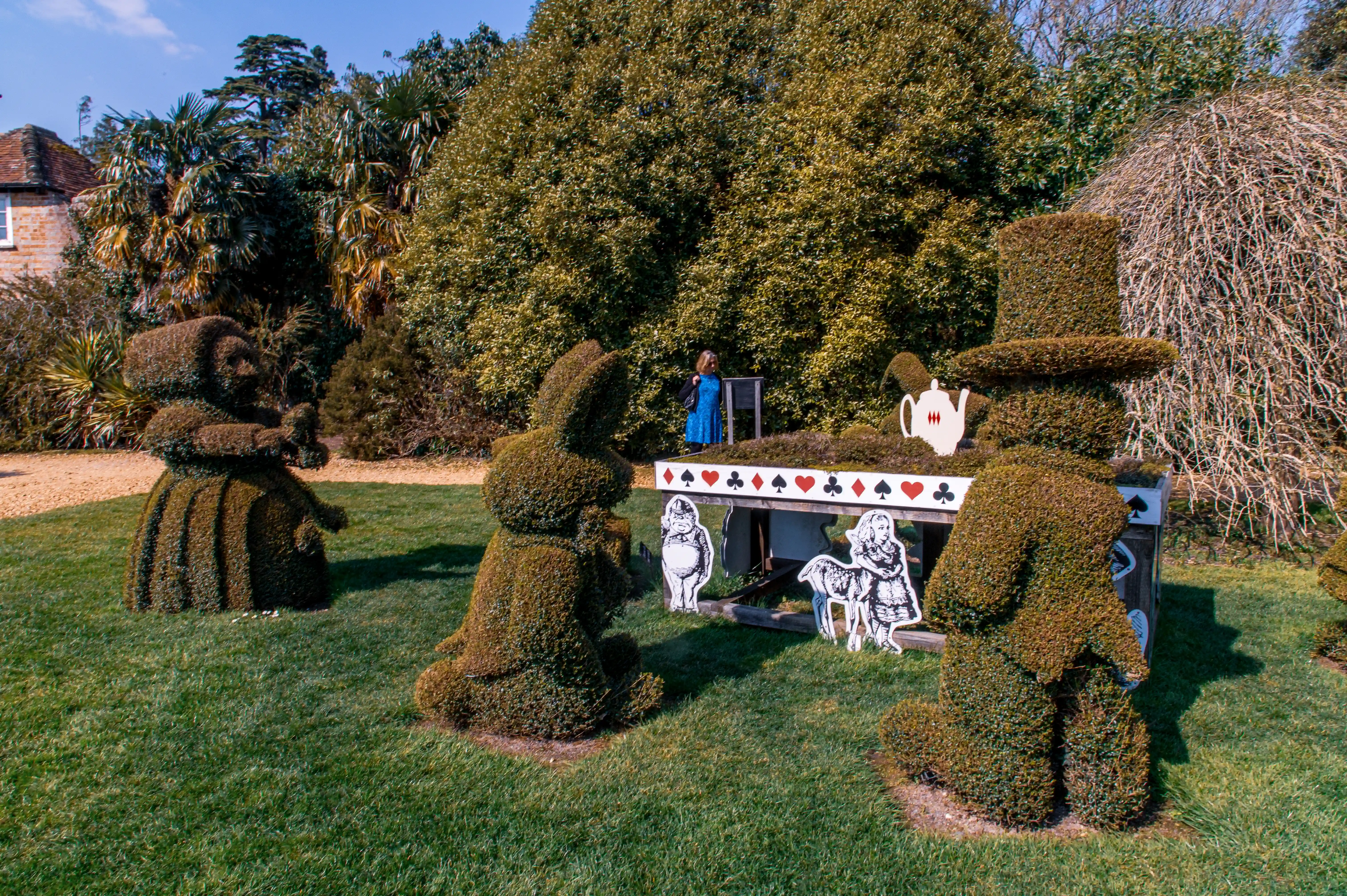 The image depicts a topiary garden scene inspired by 'Alice in Wonderland.' It features hedges sculpted into characters such as the Cheshire Cat, the Mad Hatter's teacup, and Alice herself. Additionally, there are cutouts of characters like the Cheshire Cat, Alice, and the March Hare, along with a table adorned with playing cards. The scene is set in a lush garden with various plants and trees in the background.