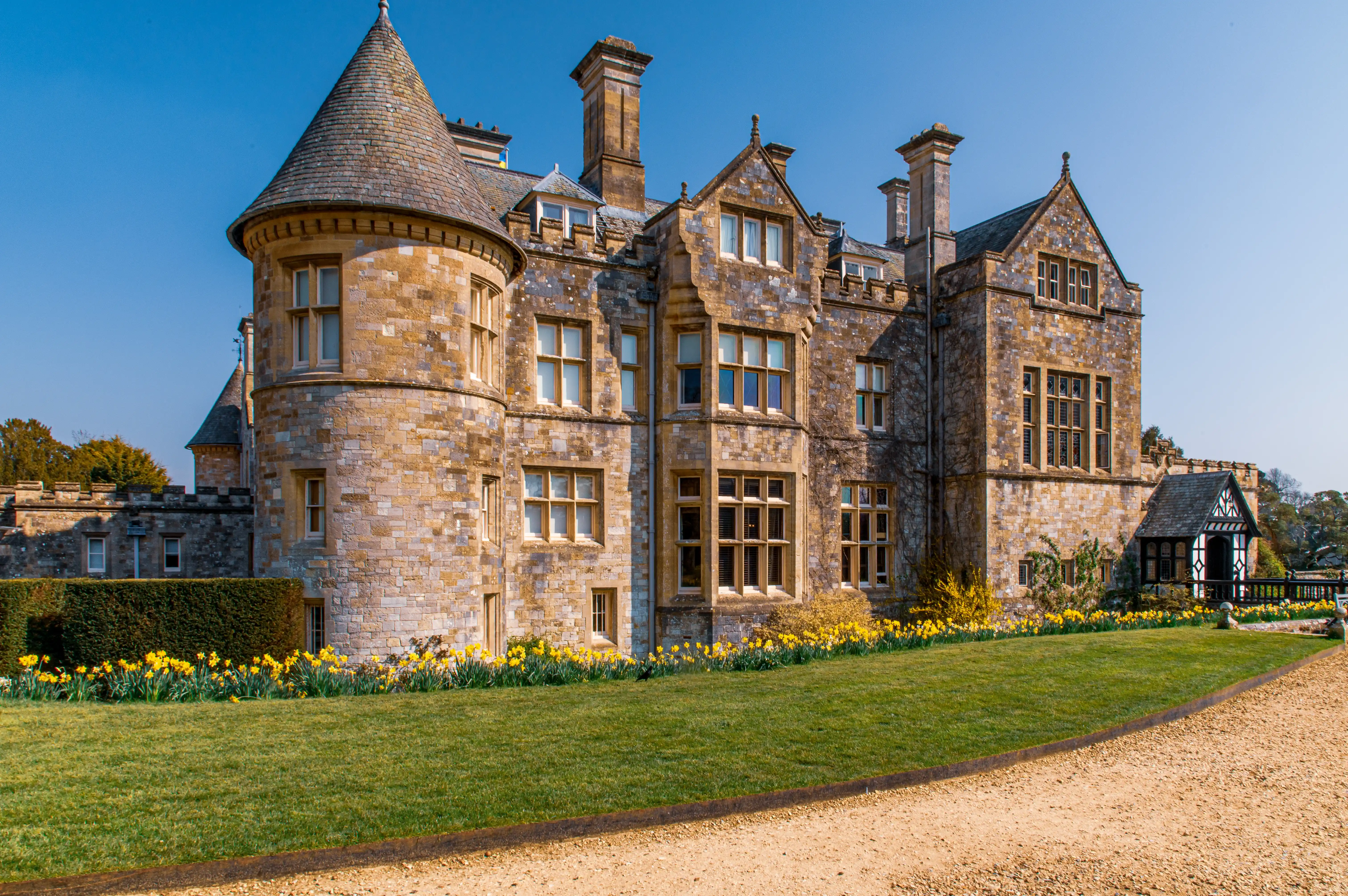 The image depicts a large, historic stone building with multiple turrets and chimneys, surrounded by a well-maintained garden with yellow flowers and a gravel pathway.