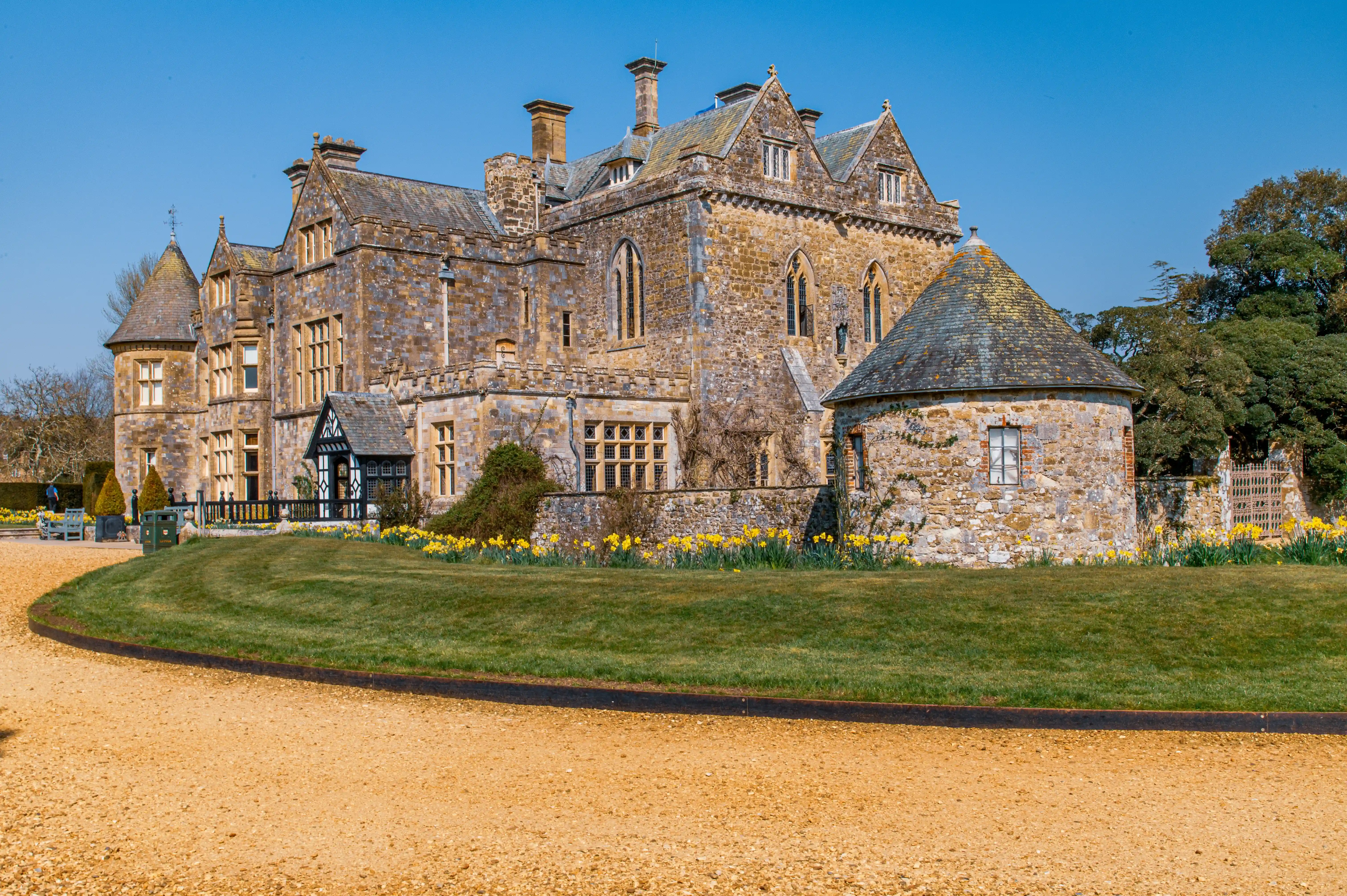 The image depicts a large, historic stone building with multiple towers and a steeply pitched roof. The structure features a combination of pointed and rounded architectural elements, with numerous windows and a central chimney. The building is surrounded by a well-maintained garden with a gravel path and vibrant yellow flowers in the foreground. The overall scene is set against a clear blue sky, suggesting a sunny day.