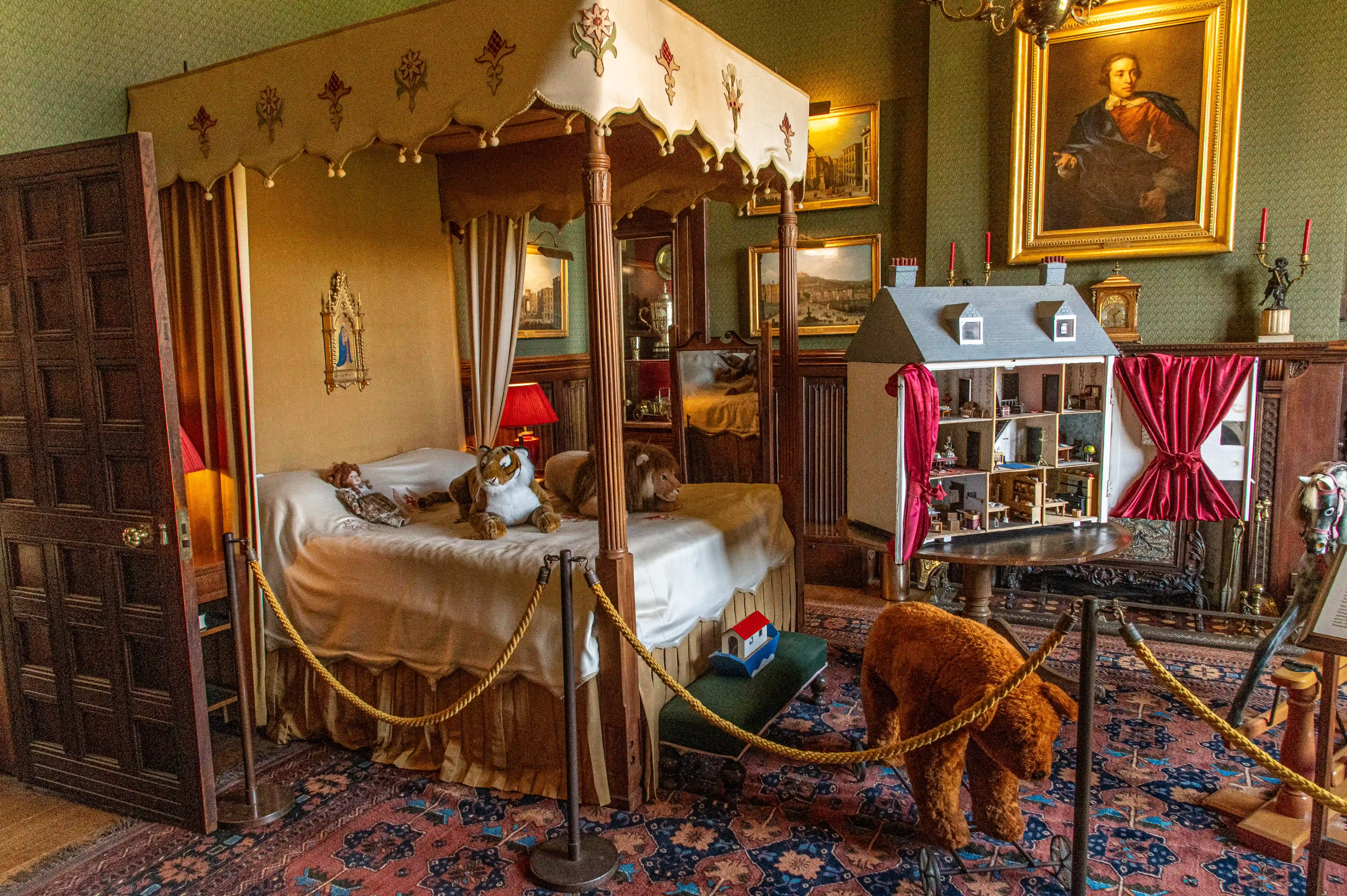 The image depicts a meticulously arranged room with a large, ornate bed adorned with stuffed animals. The room features rich, antique decor including a wooden door, framed paintings, and a dollhouse with detailed miniature furnishings. The space is cordoned off with ropes, suggesting it is part of a museum or exhibit. The overall atmosphere is one of historical elegance and careful preservation.