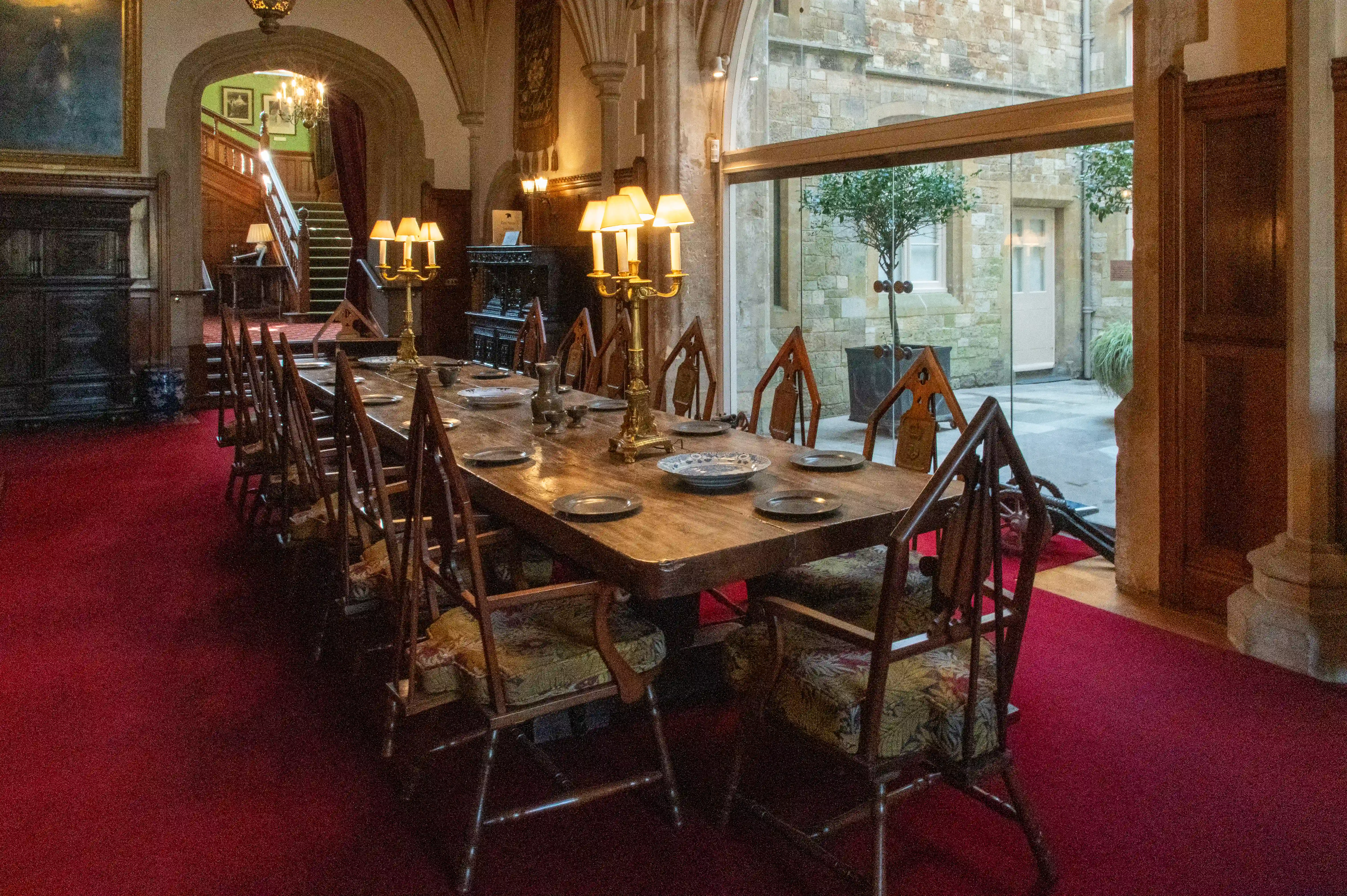 The image depicts an elegantly set dining room in what appears to be a historic or luxurious building. The room features a long wooden table with antique chairs, each set with plates and candelabras. The floor is covered with a red carpet, and the room is adorned with stone walls, large windows, and a grand staircase in the background. The architectural details, including arches and columns, suggest a medieval or Gothic style. The overall atmosphere is one of opulence and historical charm.