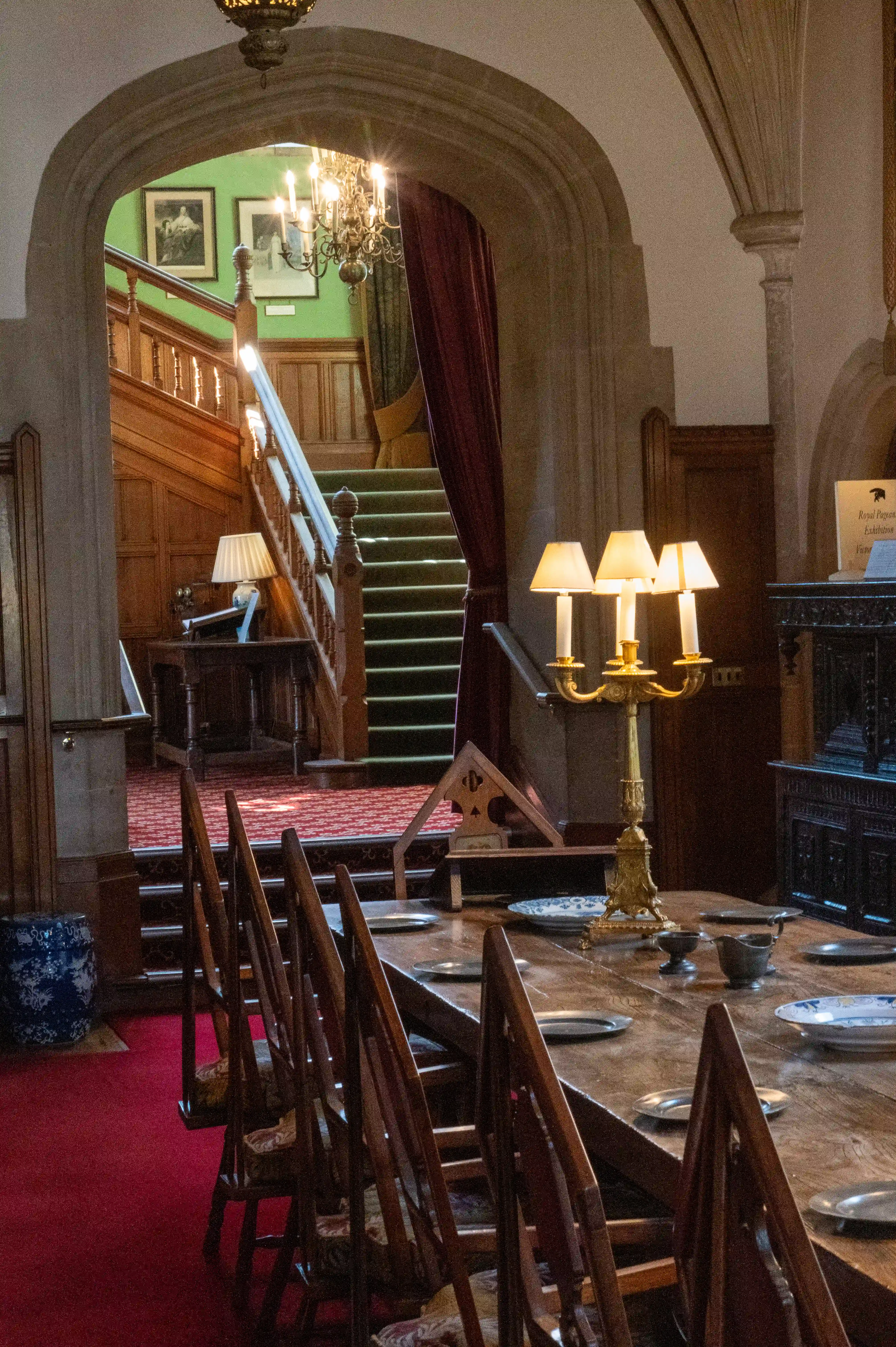 The image depicts an interior view of a grand, historic building. The focal point is a large wooden dining table set with plates and candlesticks, surrounded by high-backed wooden chairs. The room features a staircase with a wooden handrail and green carpet, leading up to a landing adorned with framed pictures. The walls and ceiling have intricate architectural details, including stone arches and a chandelier hanging from the ceiling. The overall ambiance suggests a formal dining area within a castle or manor house.