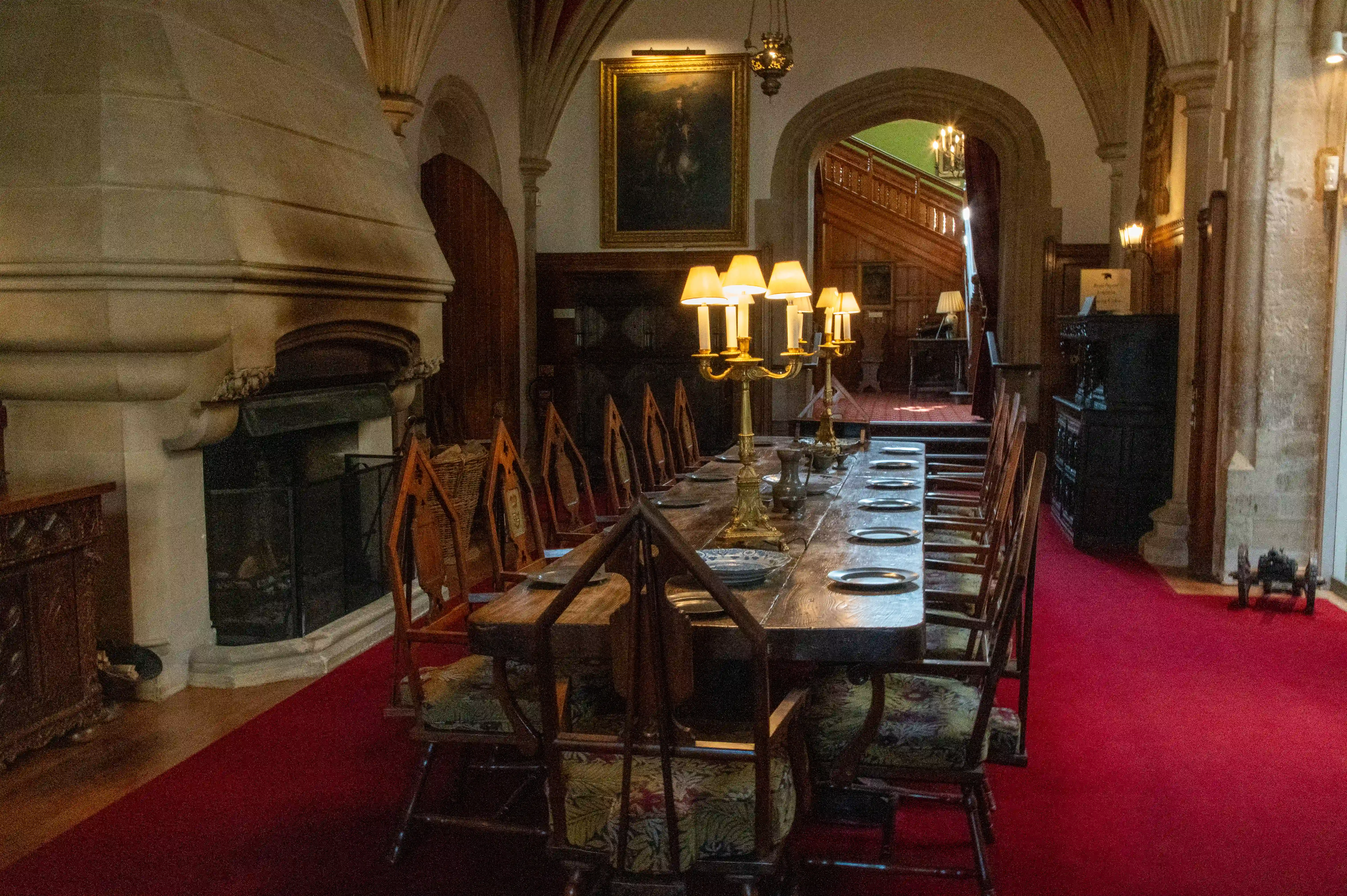 The image depicts an elegant, vintage dining room with a long wooden table set with candelabras and plates. The room features high, arched ceilings with stone walls, a grand fireplace, and a red carpet running down the center. There are wooden chairs with cushioned seats arranged around the table, and the room is illuminated by both chandeliers and wall-mounted lamps. The overall ambiance suggests a historical or formal setting, possibly within a castle or a grand manor.
