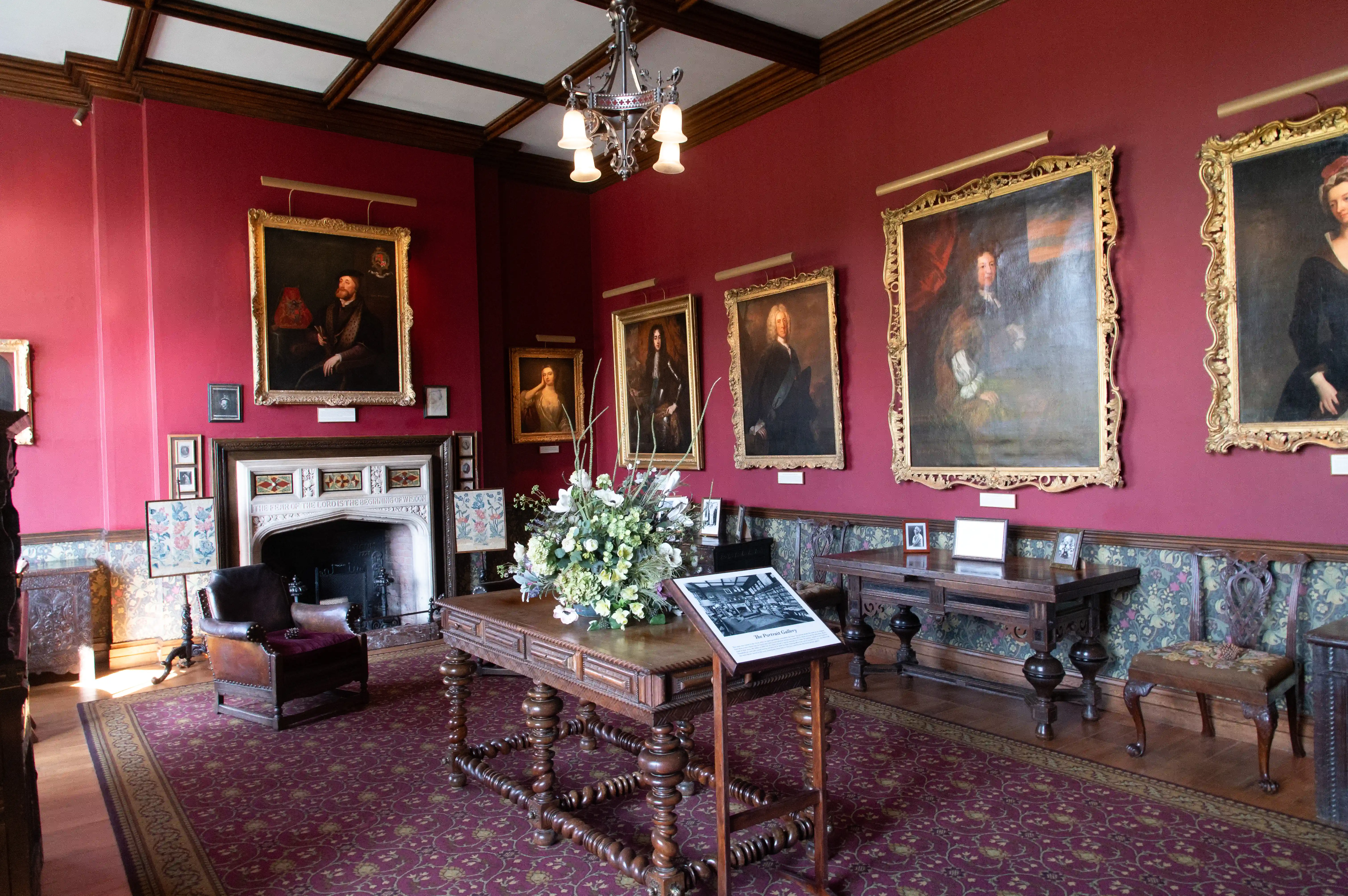 The image depicts a richly decorated room with deep red walls and ornate wooden furniture. The room features several large, gold-framed portraits of historical figures, a central table with a flower arrangement, and a fireplace with intricate detailing. There are also informational displays and smaller framed pictures on the walls and furniture, contributing to the room's historical and elegant ambiance.