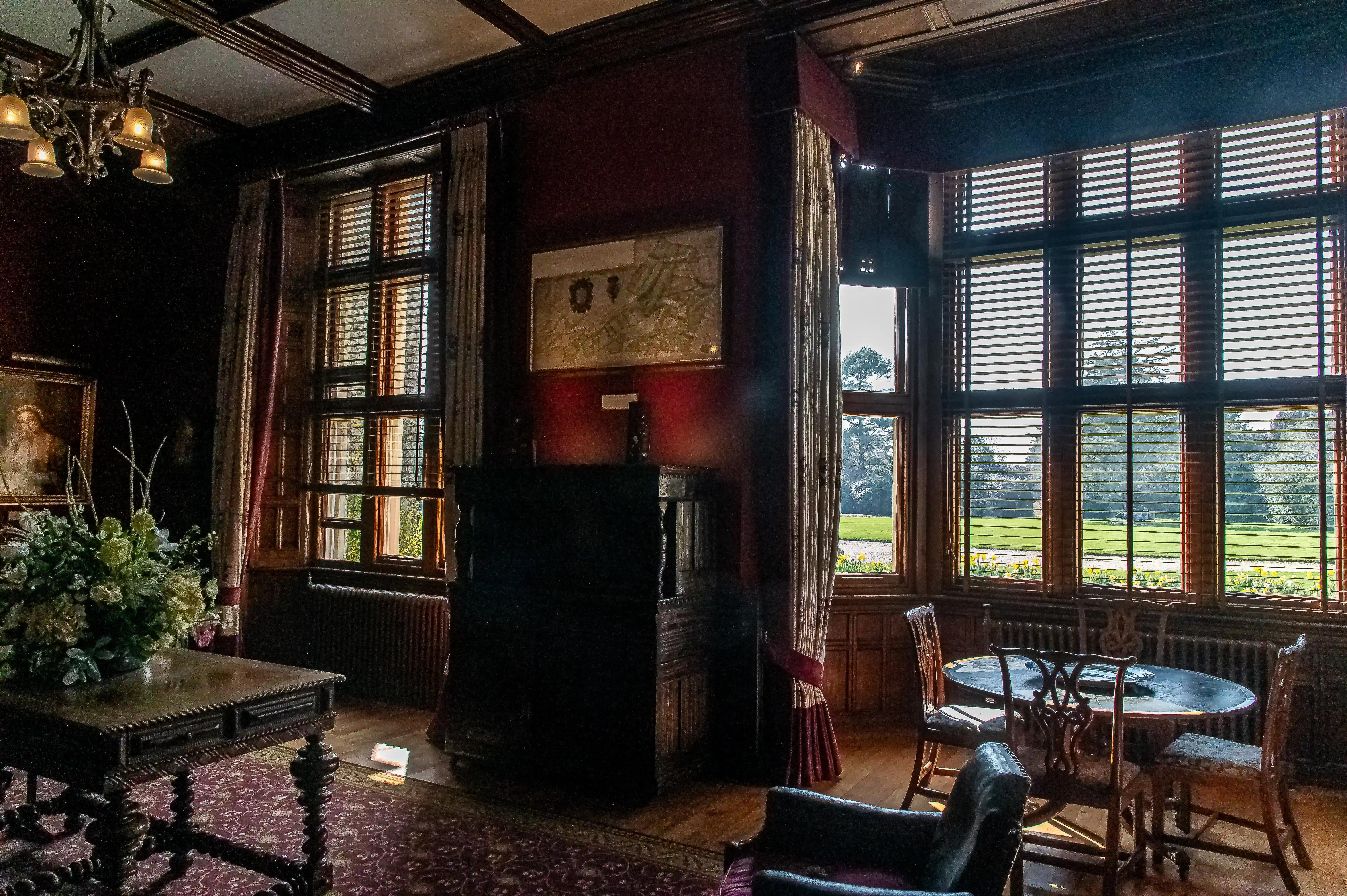 The image depicts a classic, dark-themed room with large windows allowing natural light to stream in. The room features antique furniture, including a large wooden table with intricate carvings, a smaller round table with wooden chairs, and a fireplace. There is a chandelier hanging from the ceiling, and the walls are adorned with a framed map and a painting. The room also includes a large flower arrangement on the main table, and the windows are dressed with heavy curtains and wooden blinds. Outside the windows, a well-maintained lawn and garden are visible.