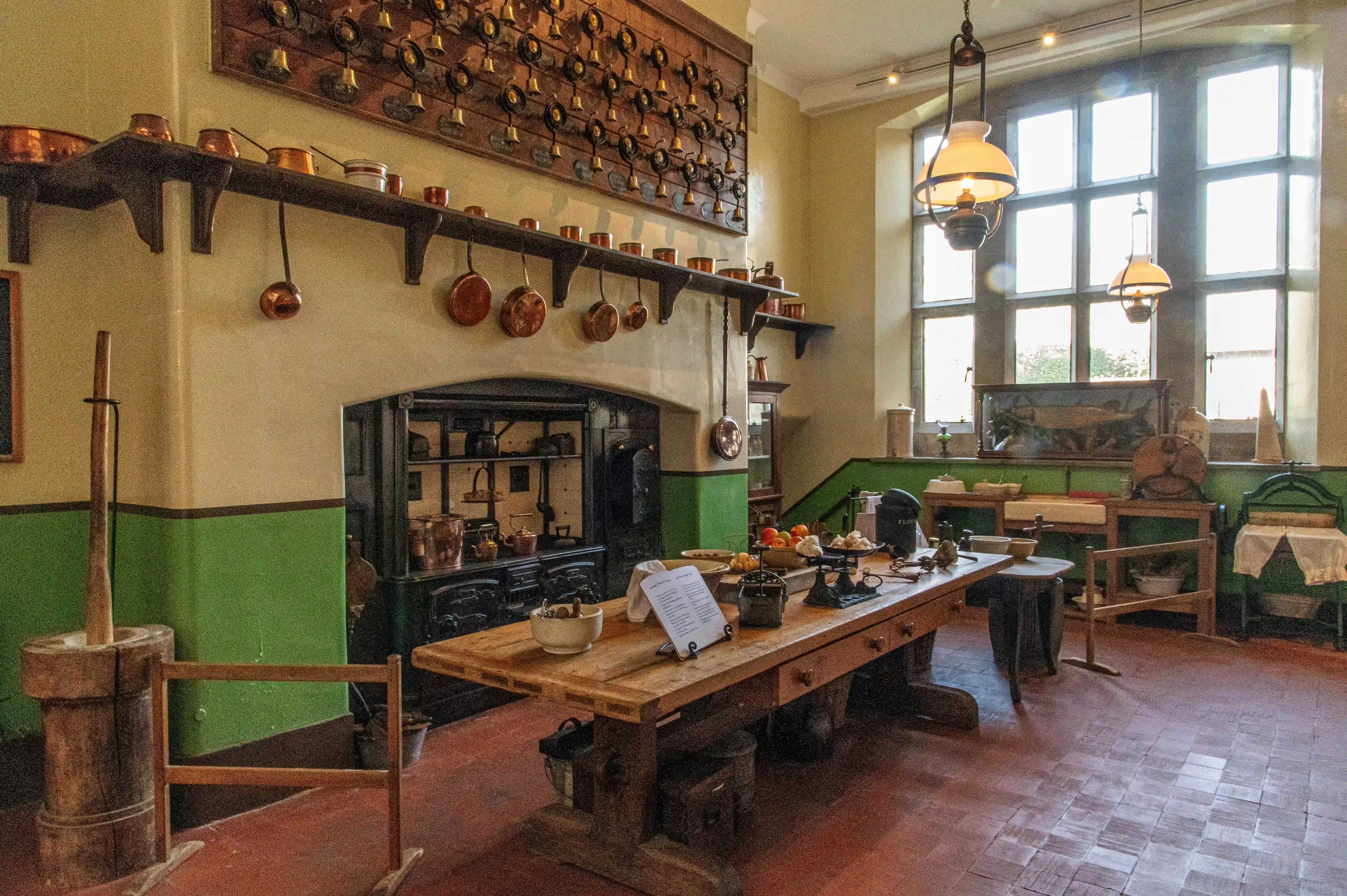 The image depicts a traditional kitchen with various vintage cooking utensils and appliances. The room features a large wooden table in the center, surrounded by other wooden furniture. Copper pots and pans are hanging on the walls, and there is a large, old-fashioned stove with multiple compartments. The kitchen has a rustic and historical ambiance, with green and beige walls, large windows allowing natural light, and several hanging lamps providing illumination.