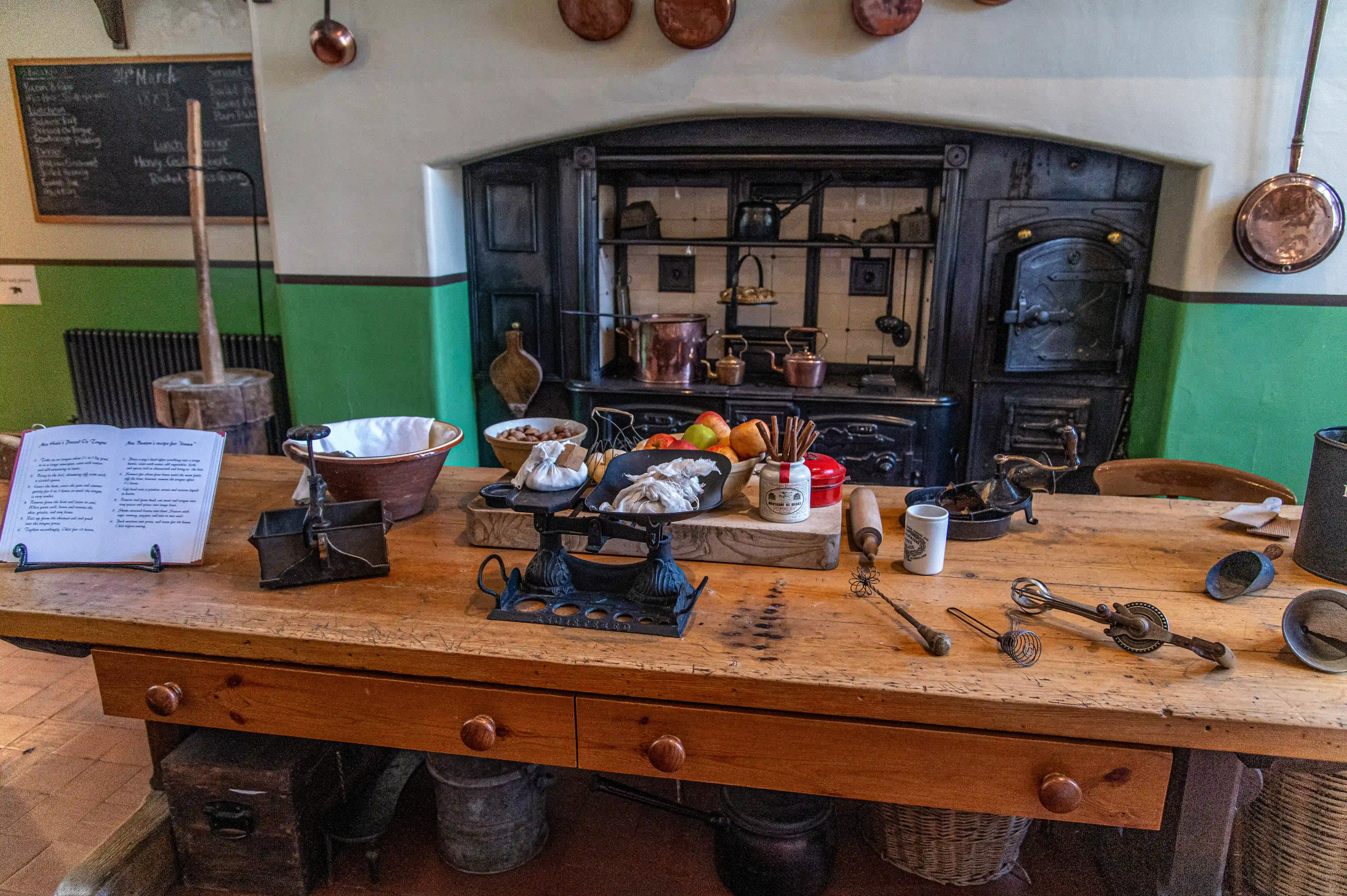 The image depicts a vintage kitchen setting with a wooden worktable cluttered with various old-fashioned kitchen tools and ingredients. The background features a large, antique stove with copper pots and pans hanging around it. The walls are painted green and white, and there are chalkboards with handwritten notes. The overall scene evokes a historical or rustic atmosphere, reminiscent of a bygone era of cooking.