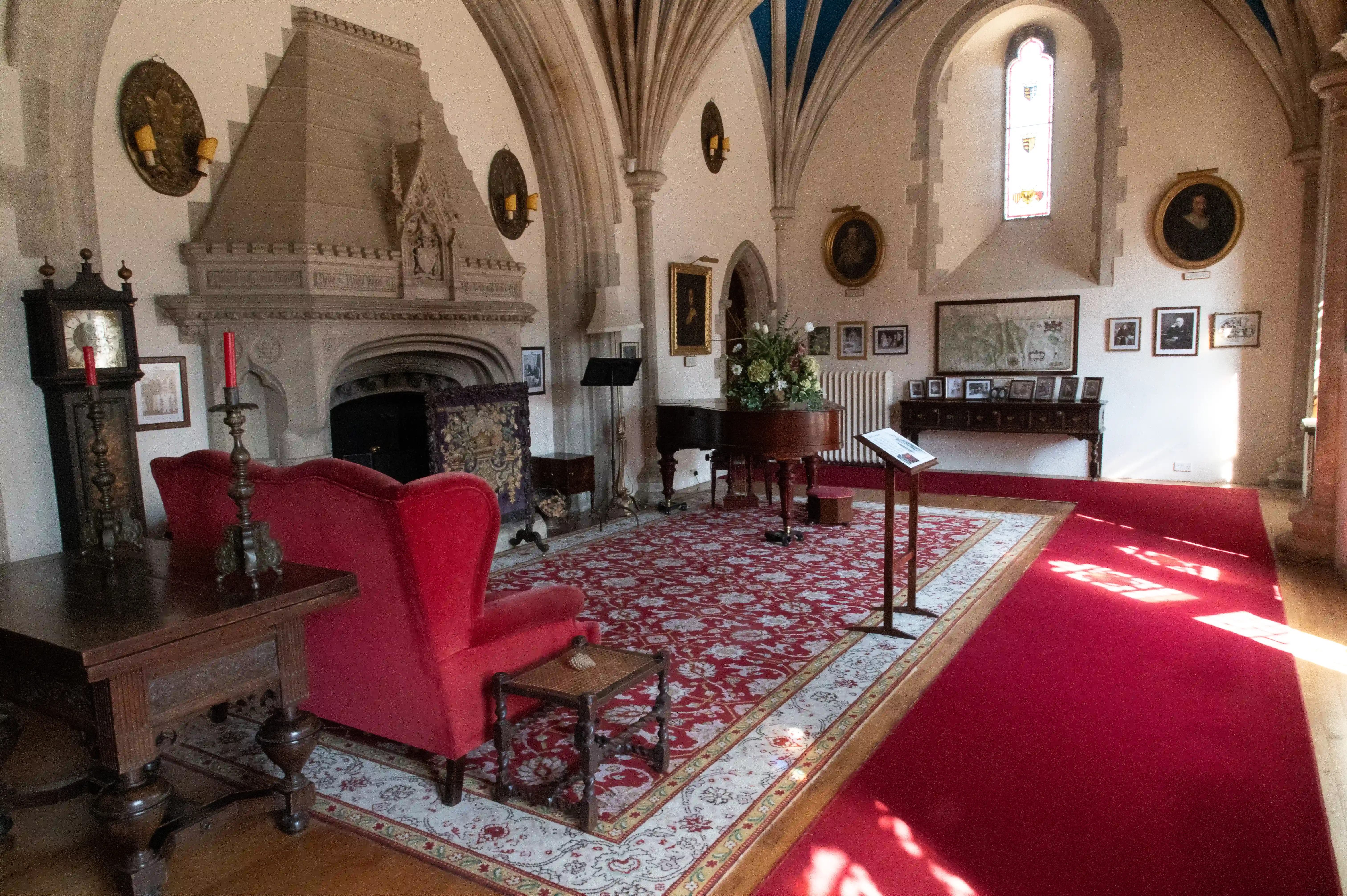 The image depicts an elegantly furnished room with a Gothic architectural style, featuring a large stone fireplace, arched windows, and intricate ceiling designs. The room is adorned with various portraits, photographs, and decorative items. The furniture includes red upholstered chairs, a wooden table, a piano, and a grandfather clock. The floor is covered with a red and cream patterned carpet, and the room is bathed in natural light from the windows.
