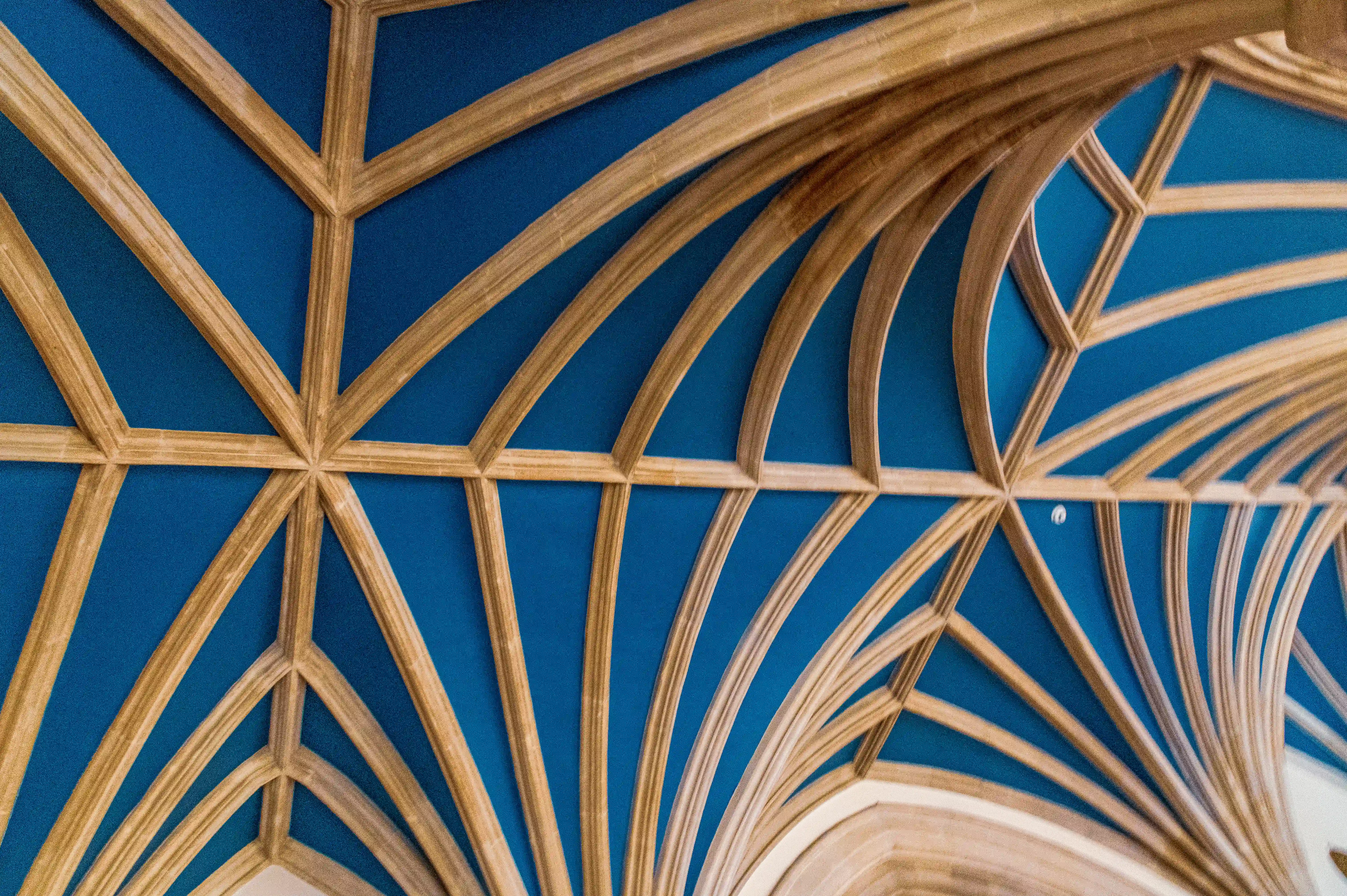 The image depicts an intricate architectural ceiling design featuring a combination of geometric patterns and ribbed vaulting. The ceiling is painted in a vibrant blue color, contrasted by golden, rib-like structures that form a complex, star-like pattern. The design showcases the intersection of these ribs, creating a visually striking and symmetrical effect. The craftsmanship highlights the detailed and ornate nature of the architecture, likely indicative of a historic or significant building