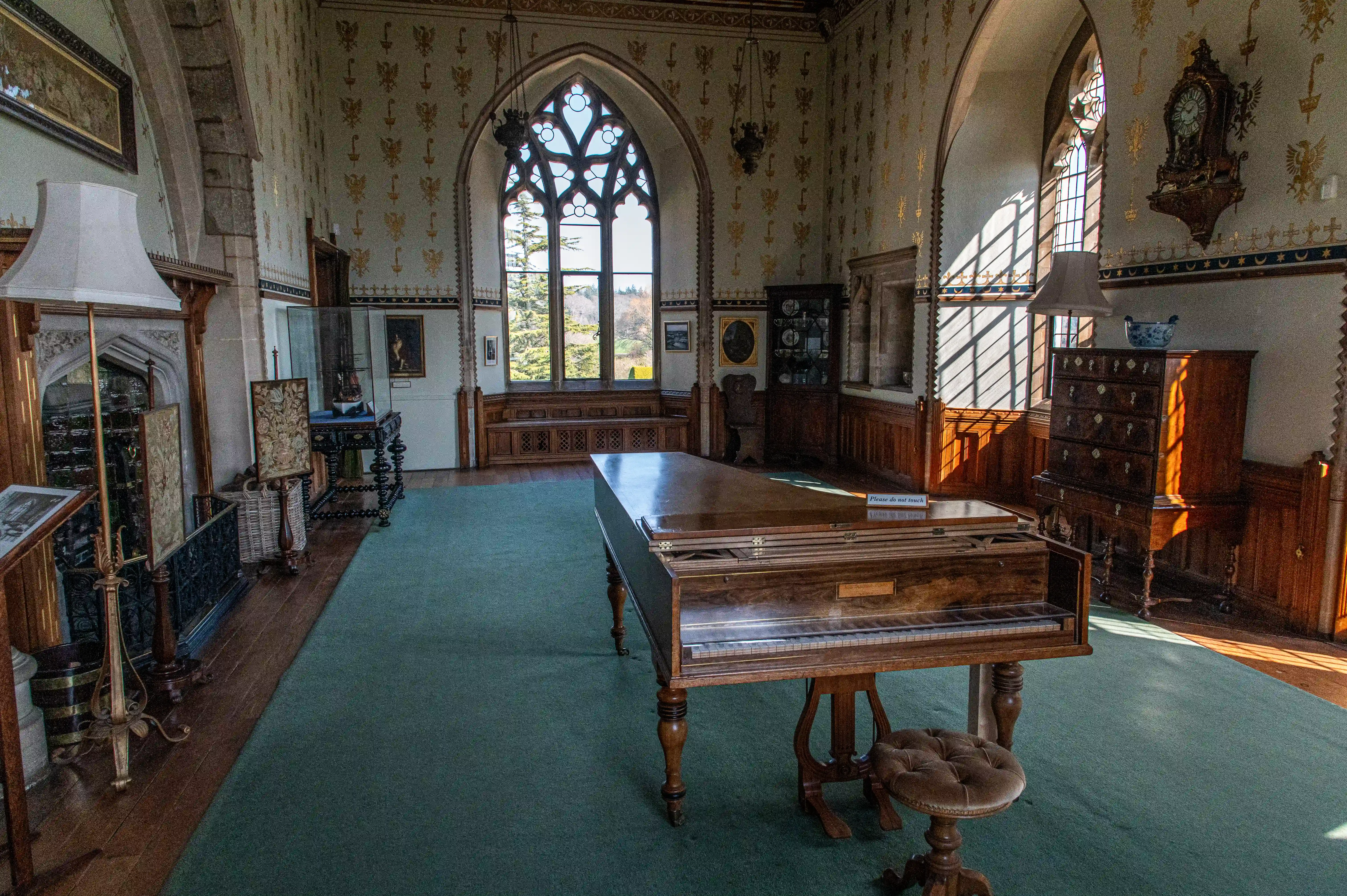 The image depicts an ornately decorated room with a grand piano as the central piece. The room features Gothic-style architecture with large arched windows, intricate wall decorations, and antique furniture. The walls are adorned with various paintings and decorative items, and the room is illuminated by natural light streaming through the windows.