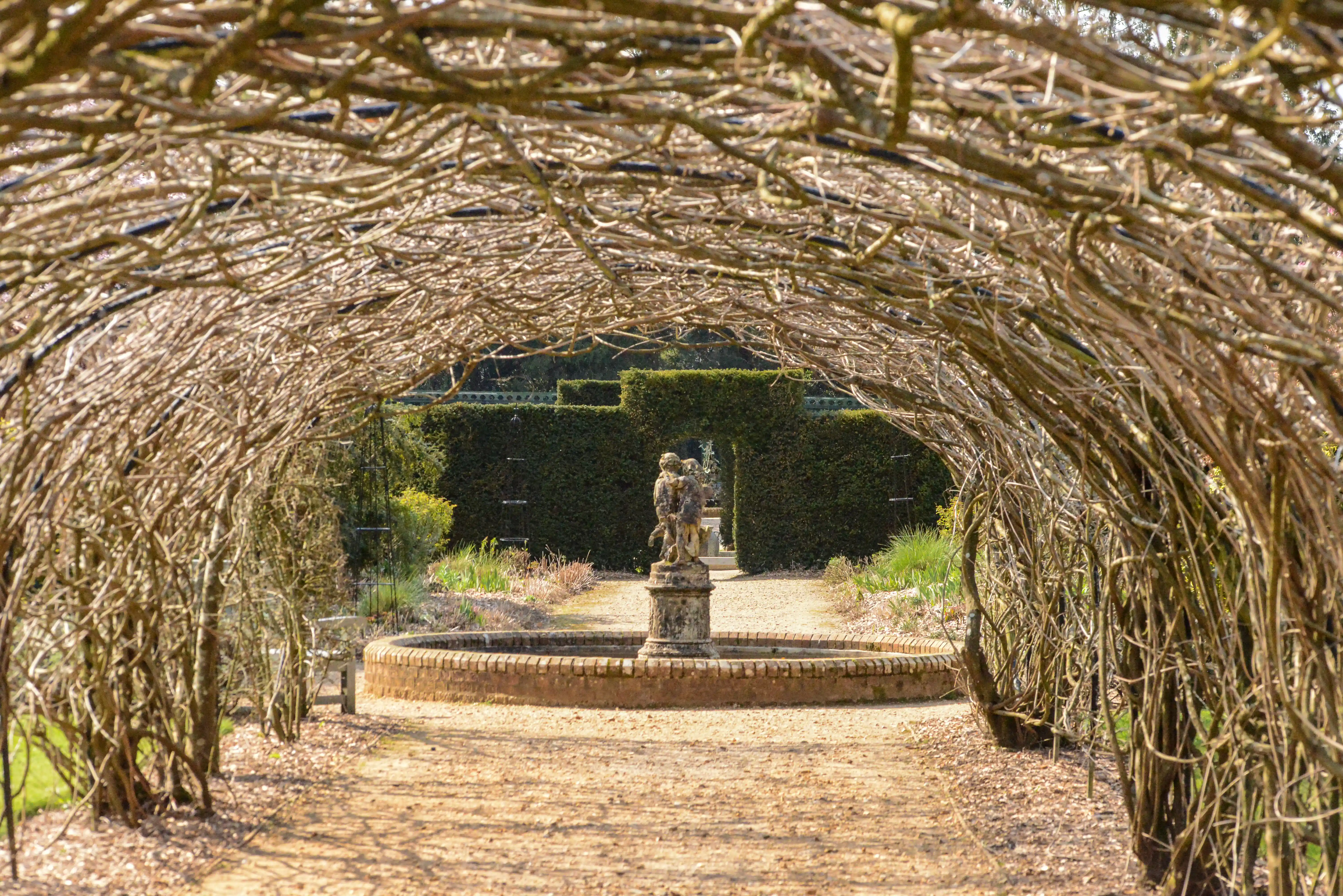 The image depicts a garden scene viewed through a tunnel-like structure made of intertwined branches. The garden features a statue on a pedestal in the center, surrounded by a circular brick border. In the background, there is a well-maintained hedge and various plants and trees. The overall atmosphere is serene and well-kept, suggesting a formal garden setting.