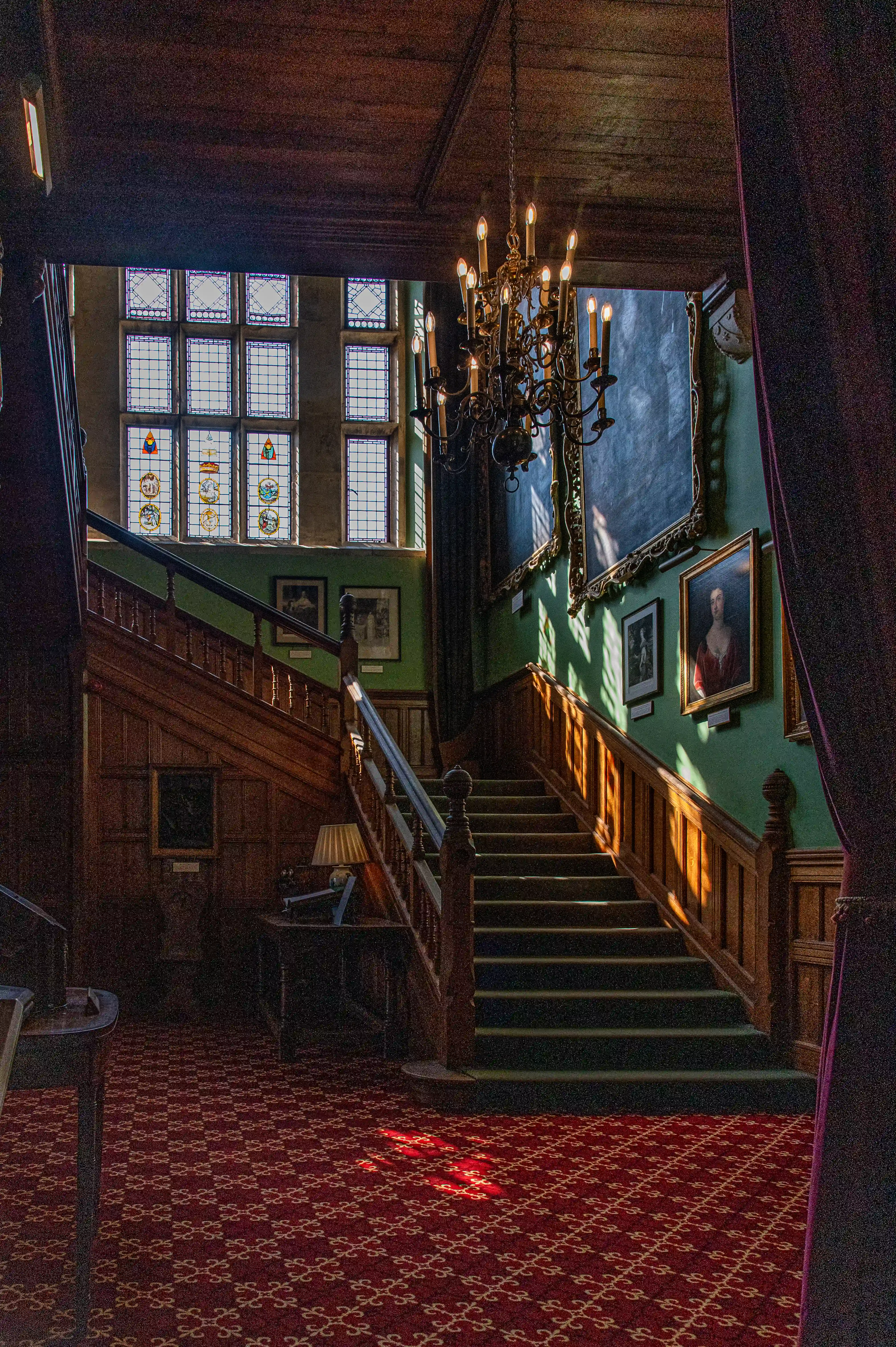 The image depicts an interior view of a grand, historic staircase with ornate wooden paneling, a chandelier, and stained glass windows. The staircase is illuminated by natural light streaming through the windows, casting a warm glow on the steps and surrounding walls. The walls are adorned with framed portraits and decorative elements, adding to the elegant and timeless atmosphere of the space.