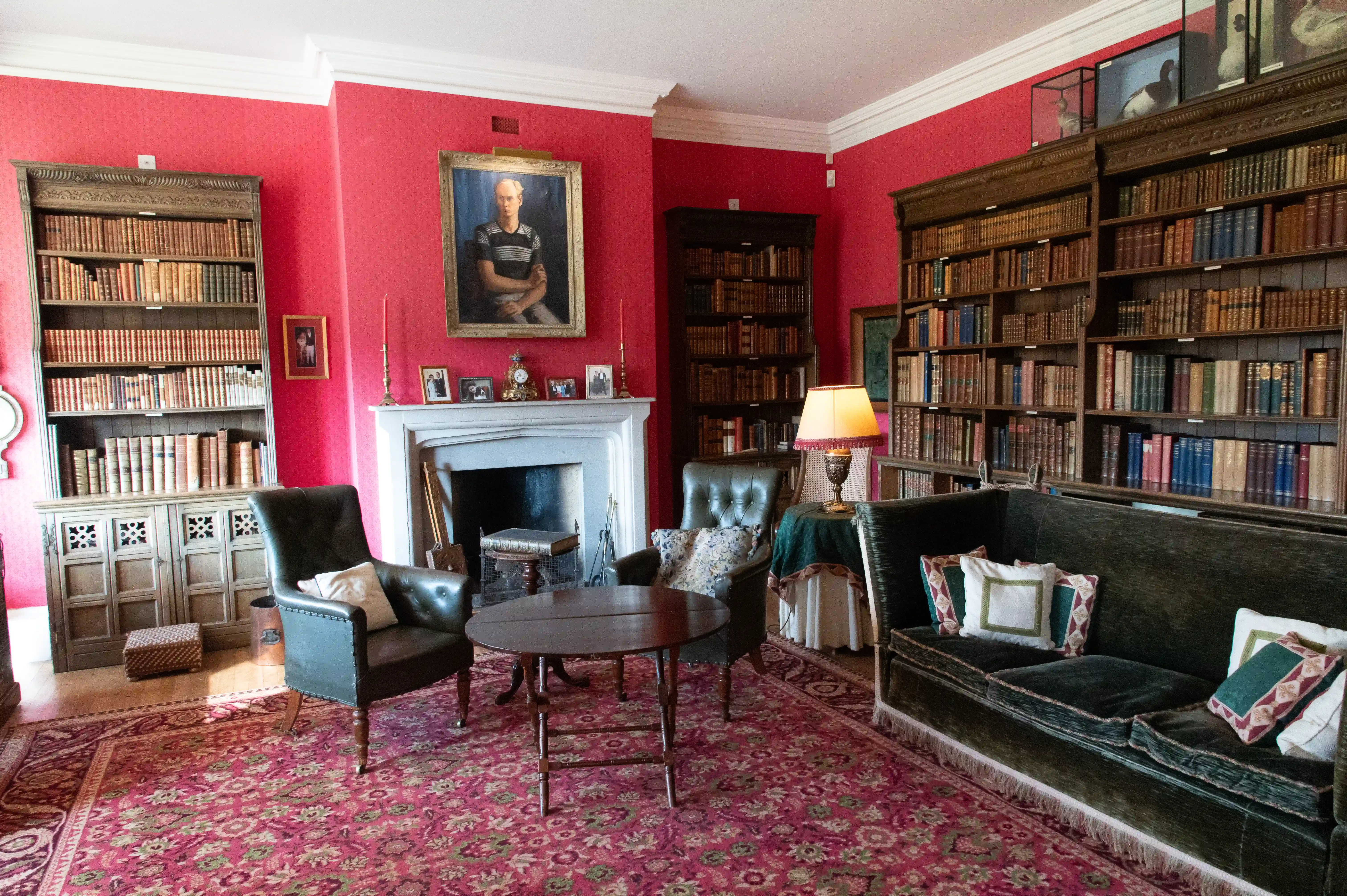 The image depicts a cozy, richly decorated library or study room with walls painted in a deep red color. The room features floor-to-ceiling bookshelves filled with books, a fireplace with a portrait above it, comfortable leather furniture including armchairs and a sofa, and a patterned rug covering the floor. There are also decorative items such as lamps, cushions, and small tables, creating an inviting and warm atmosphere.