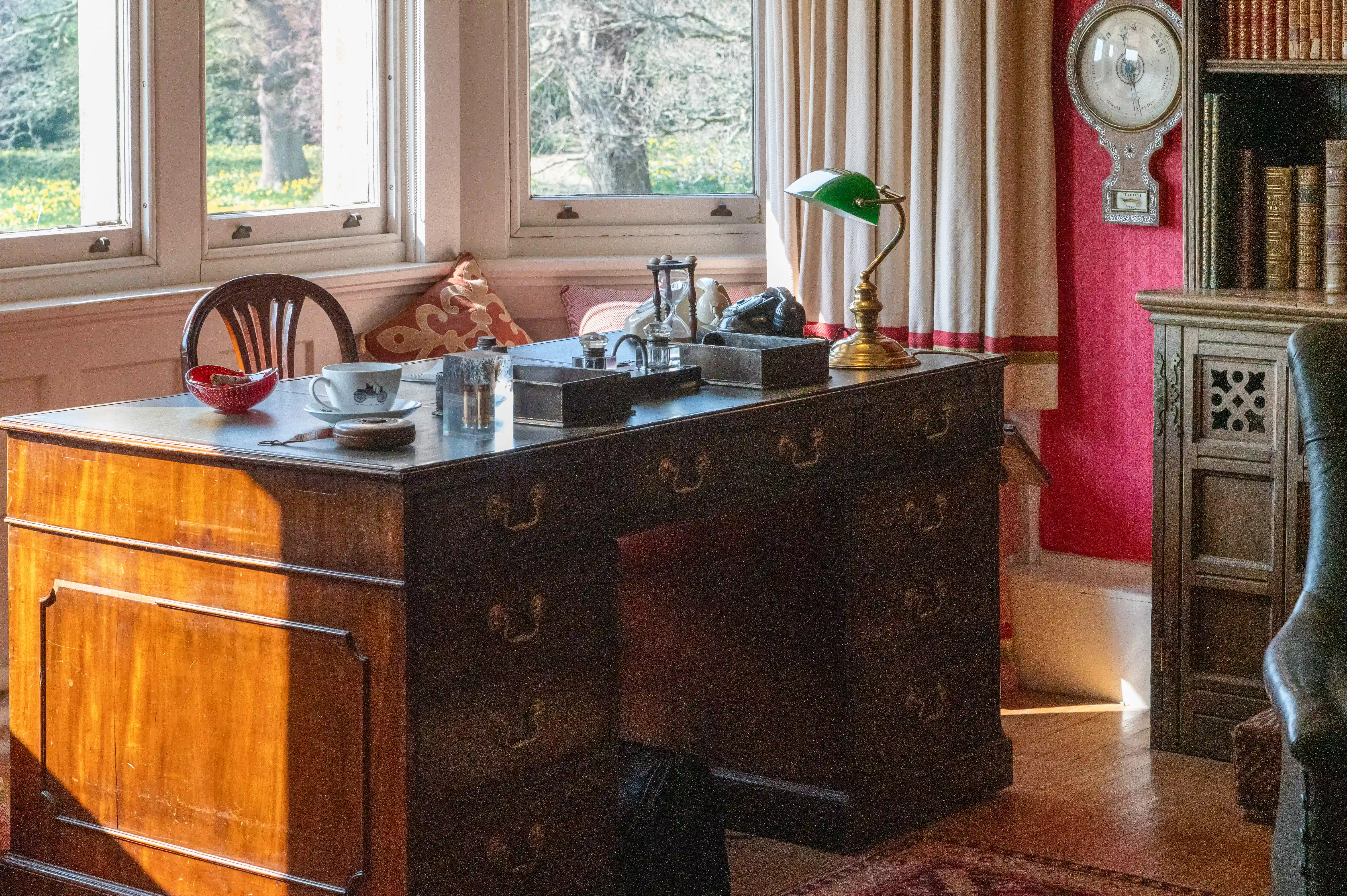 The image depicts a vintage-style study or office room with a wooden desk positioned near a window. The desk has several items on it, including a green desk lamp, an hourglass, a typewriter, a brass lamp, a telephone, and various other small objects. There is also a red basket and a cup and saucer on the desk. The room features wooden furniture, including a chair and a cabinet, with a patterned rug on the floor. The overall ambiance suggests a cozy, well-lit space suitable for reading, writing, or other desk work.