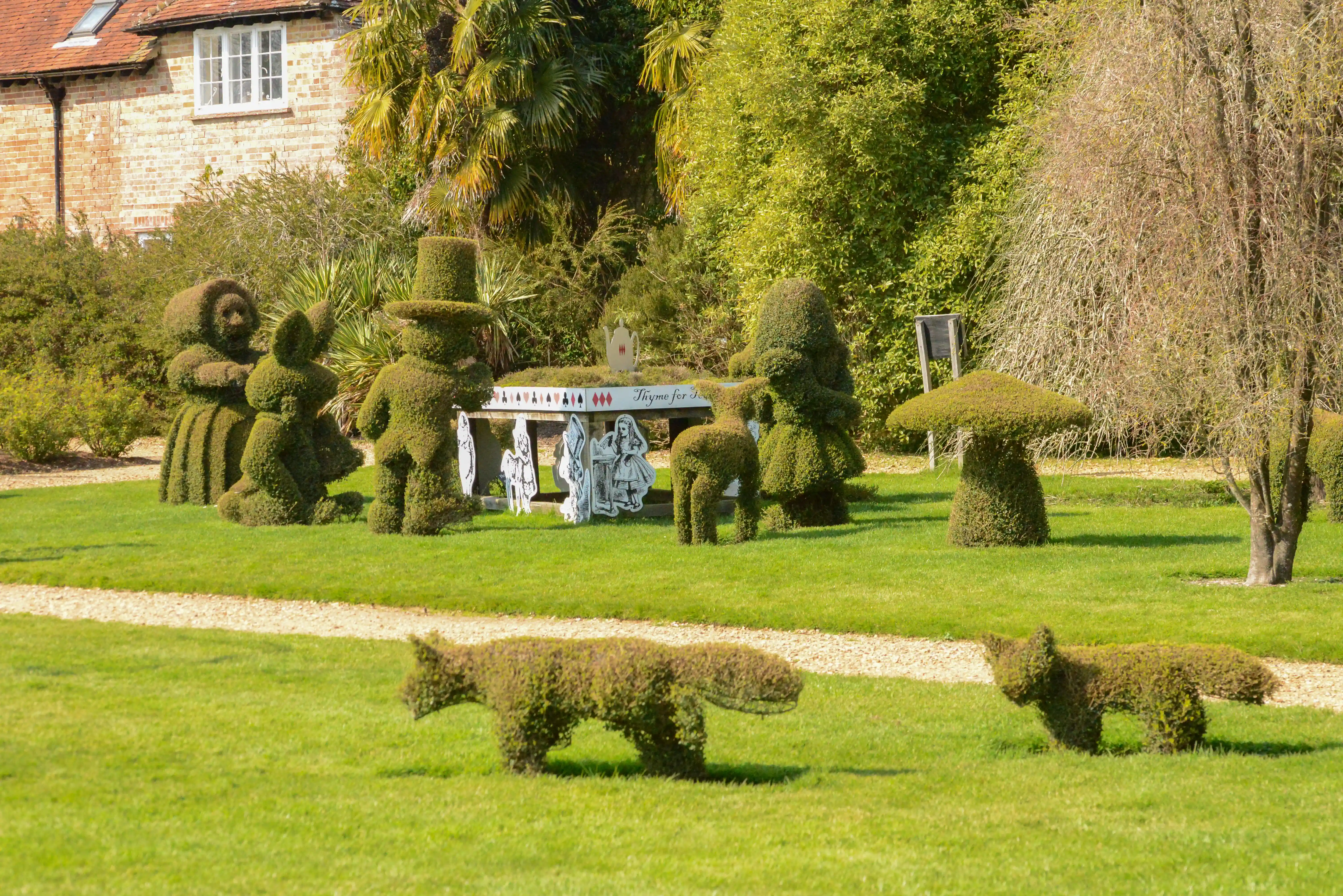The image depicts a topiary garden featuring hedges sculpted into various shapes, including characters from 'Alice in Wonderland' and animals. The garden is well-maintained with green lawns and gravel paths. In the background, there is a brick building with a tiled roof, partially obscured by trees and shrubs. The scene is set on a sunny day, with clear skies and lush greenery.