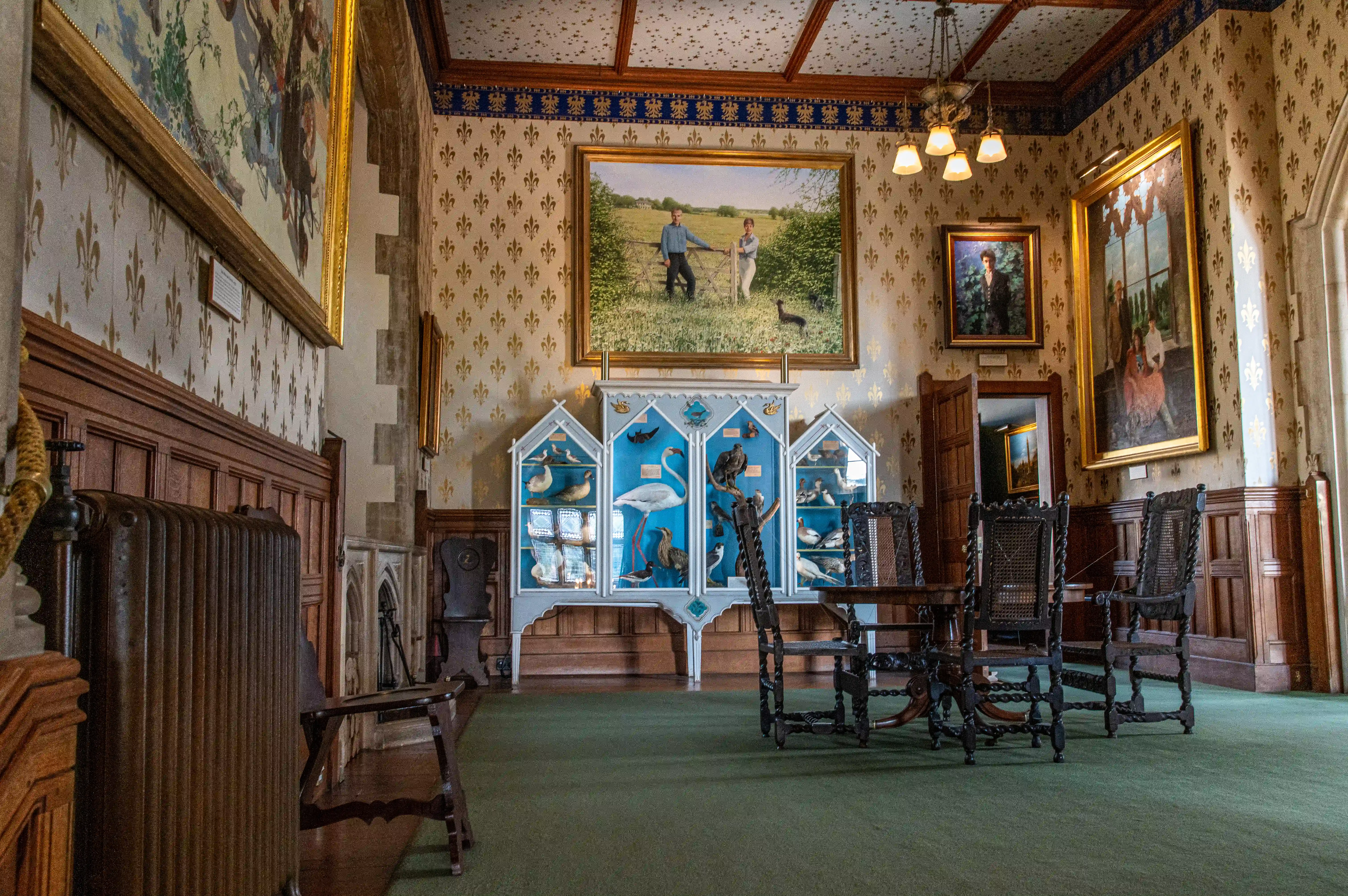 The image depicts an ornately decorated room with antique furniture and bird displays. The walls are adorned with intricate wallpaper and several framed paintings, including a large landscape painting and portraits. The room features wooden paneling, a green carpet, and a collection of antique chairs and benches. There is a display case with various birds, and the ceiling has a decorative pattern. The overall ambiance suggests a historical or museum setting.