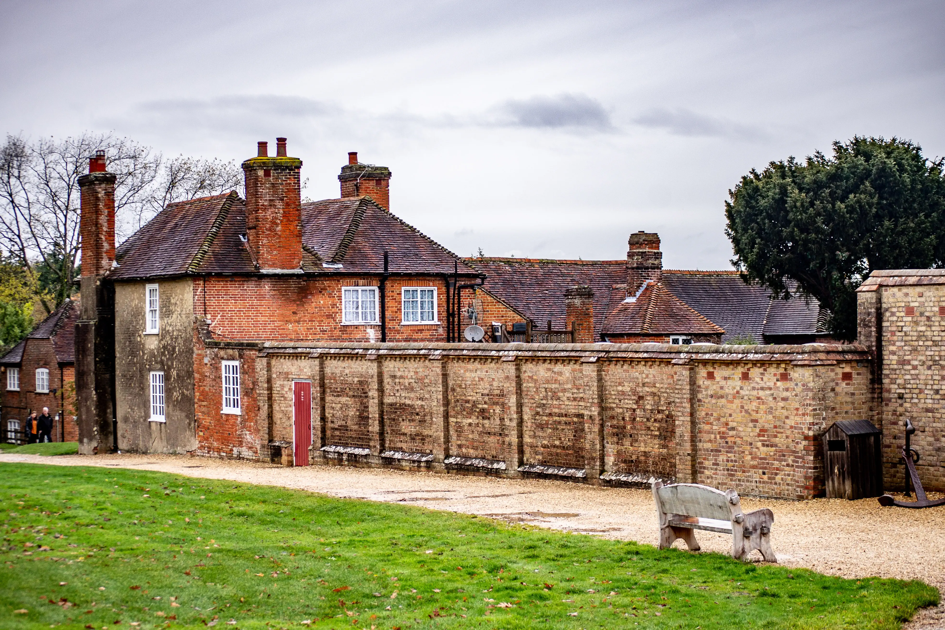 The image depicts an old, rustic building with a brick exterior and a tiled roof, surrounded by a stone wall. The building has multiple chimneys and small windows, giving it a historic appearance. The area in front of the building is a grassy lawn with a gravel path and a wooden bench. The scene is overcast, with a cloudy sky and bare trees, suggesting it might be late autumn or winter.