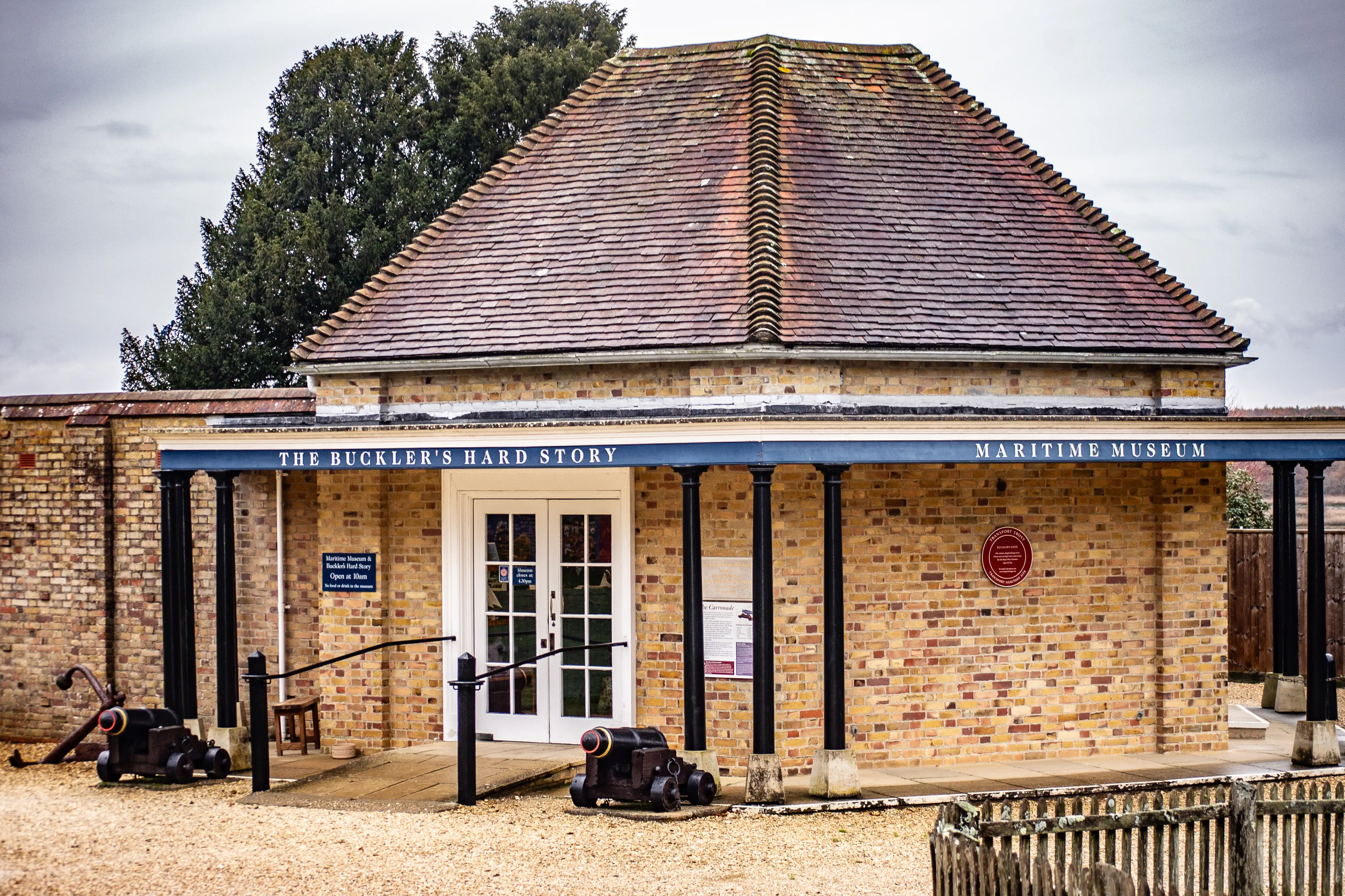 The image depicts the entrance to the 'Buckler's Hard Story' exhibit at a Maritime Museum. The building features a tiled roof and brick walls, with a sign above the entrance indicating the exhibit's name. There are several black cannons displayed in front of the building, and the entrance is accessible via a ramp and stairs. Informational signs are posted near the entrance, and the overall setting appears to be part of a historical or educational site focused on maritime history.