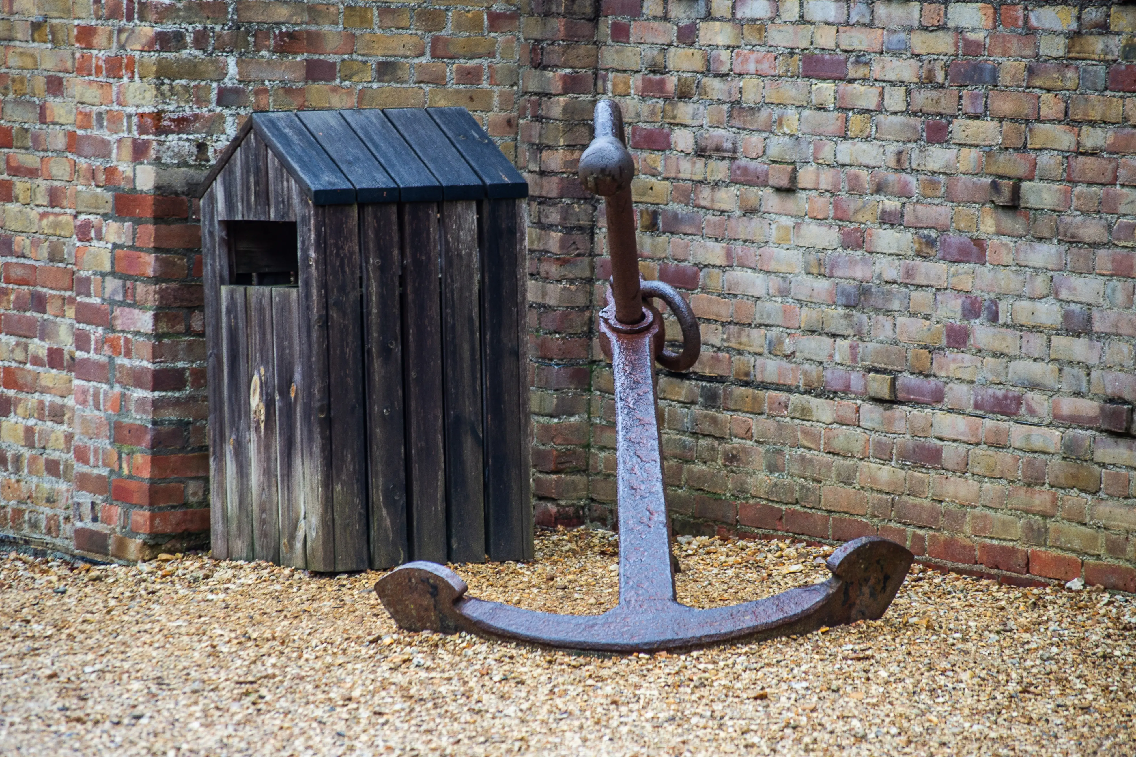 The image shows a rusted anchor and a wooden structure resembling a small, old-fashioned outhouse or shed against a brick wall background with wood chip ground cover.