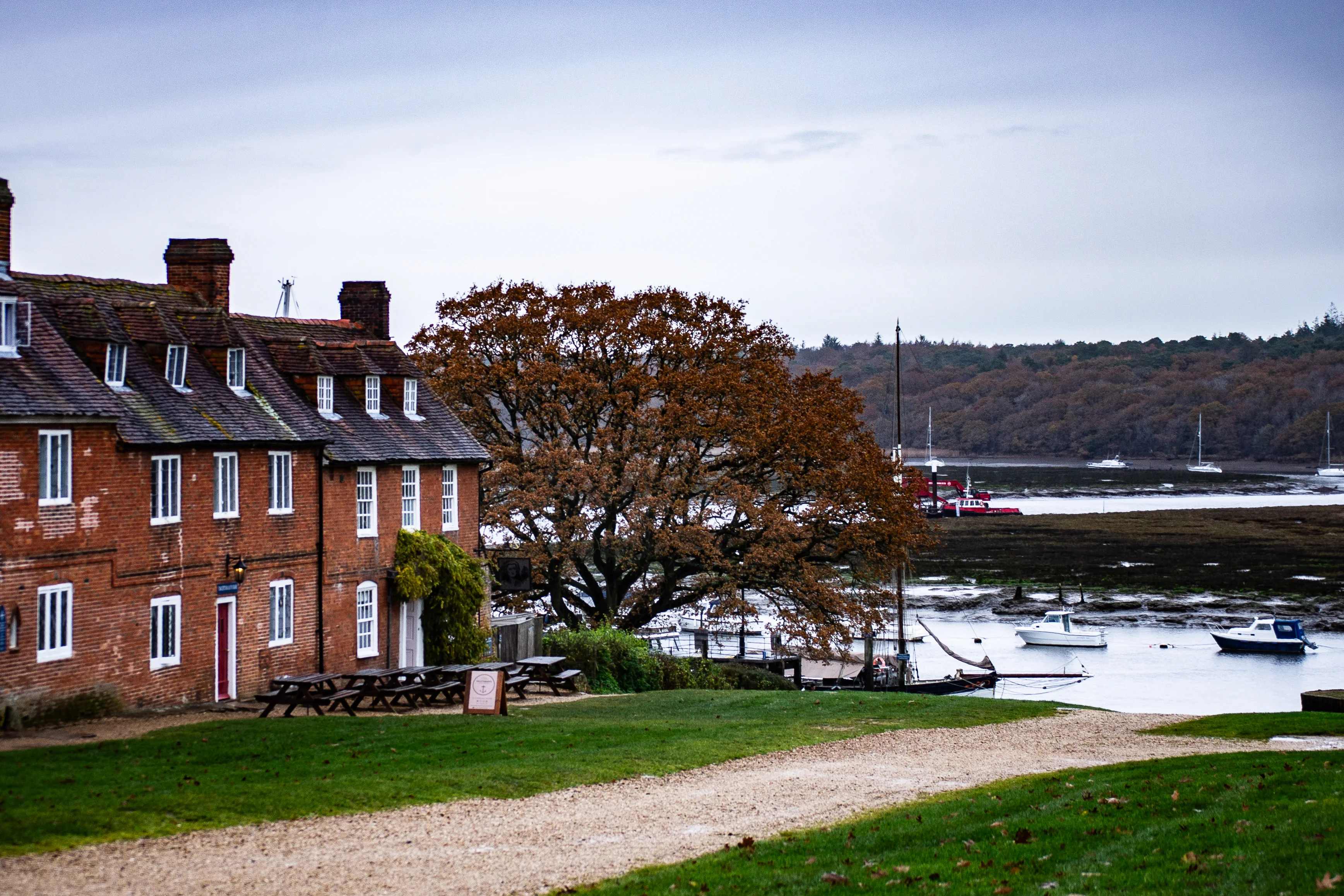 The image depicts a serene waterfront scene with a large brick house featuring white-framed windows and a tiled roof. In front of the house, there is a grassy area with picnic tables and a sign. A large tree with autumn foliage stands near the water's edge, where several boats are moored. The background shows a body of water with more boats and a hilly, forested landscape under a cloudy sky.