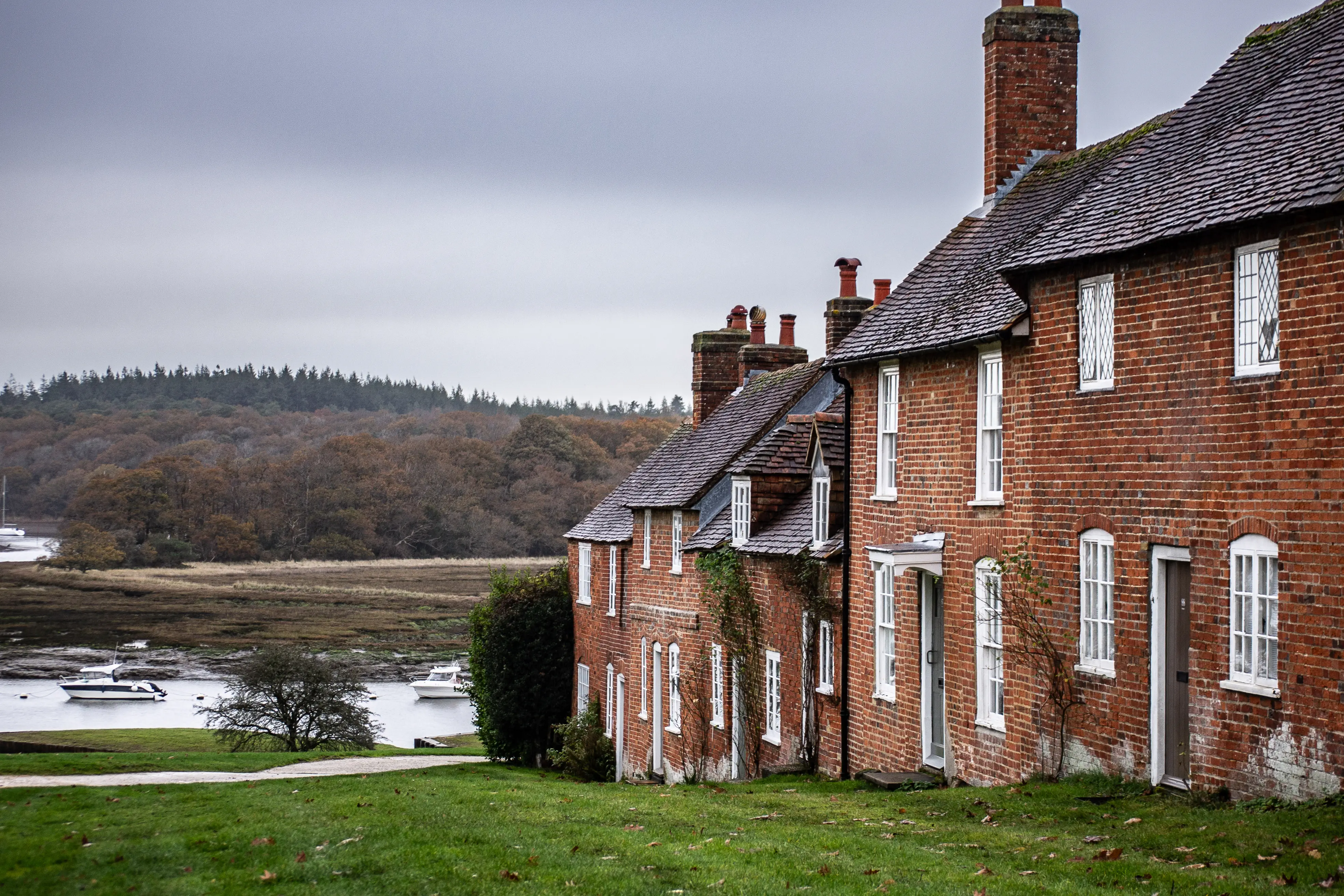 The image depicts a row of traditional brick houses with white-framed windows and doors, situated along a grassy area near a body of water. The houses have steep, tiled roofs and several chimneys. The background features a forested hillside, and the overall scene appears to be a serene, rural setting with a calm, overcast sky.